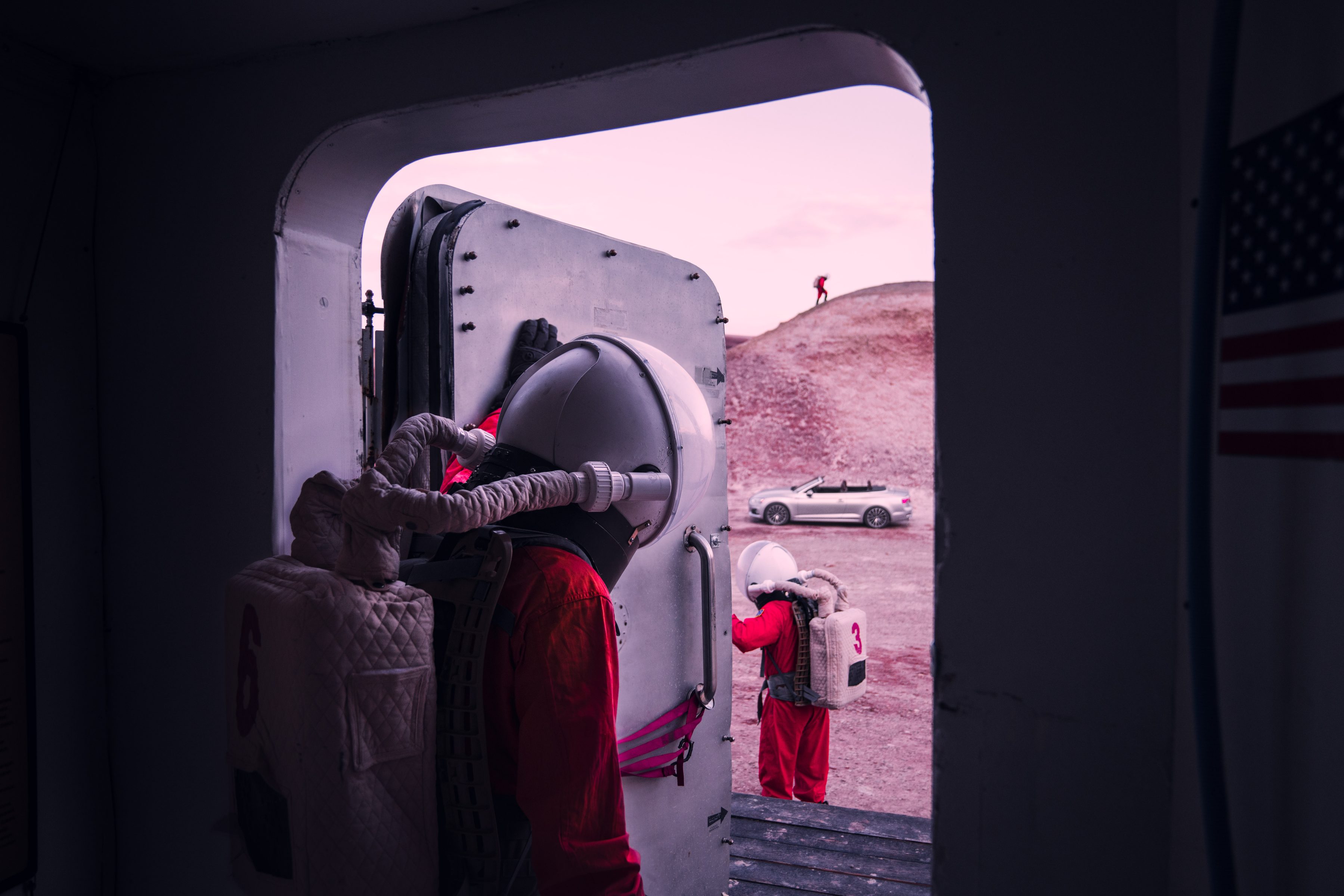 Two people in spacesuits exit a hatch onto a rocky terrain, while a car and a photographer capturing nighttime landscape photography stand outside.