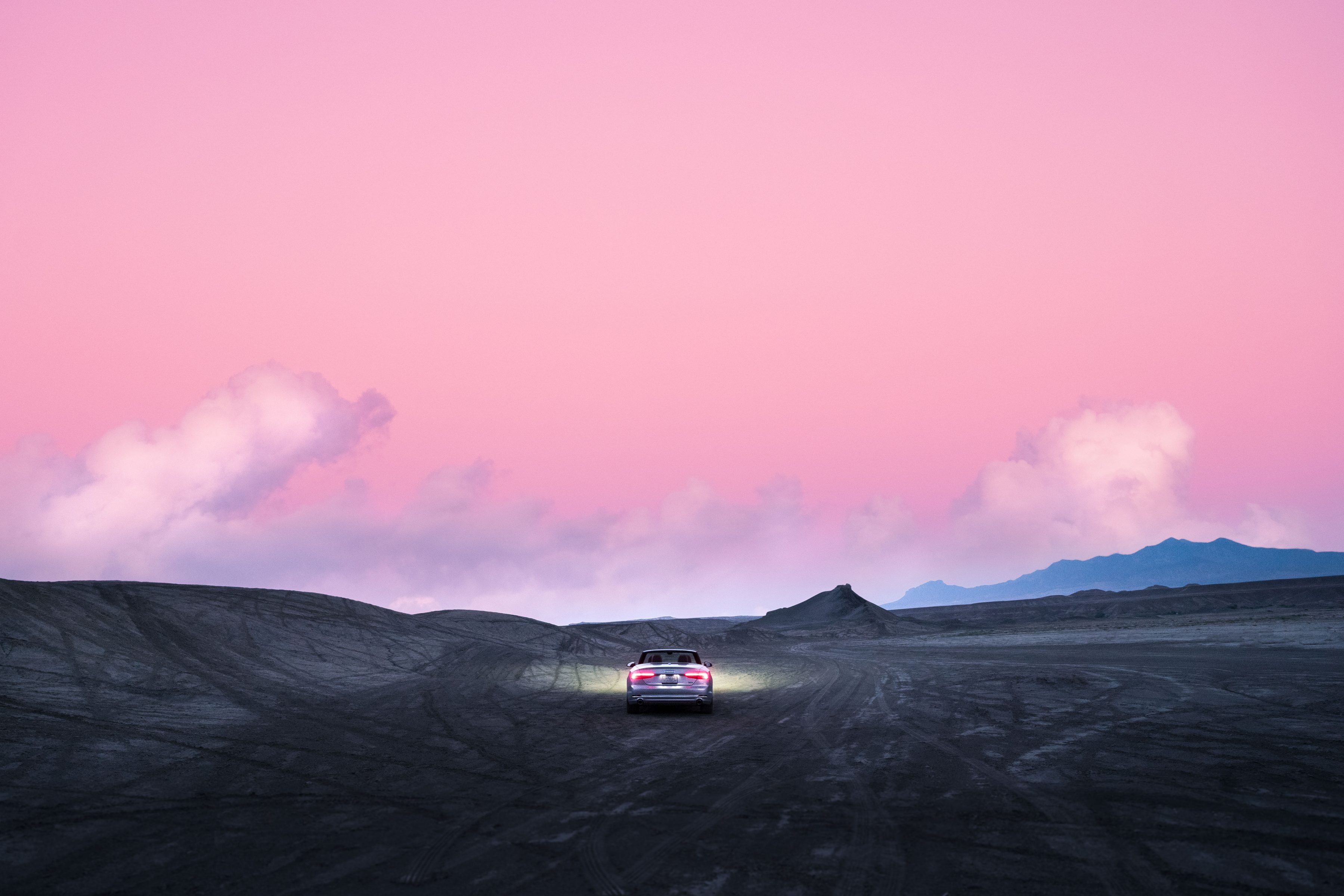 A car drives on a barren landscape at dusk under a pink sky with clouds and distant mountains, capturing the spirit of nighttime landscape photography.