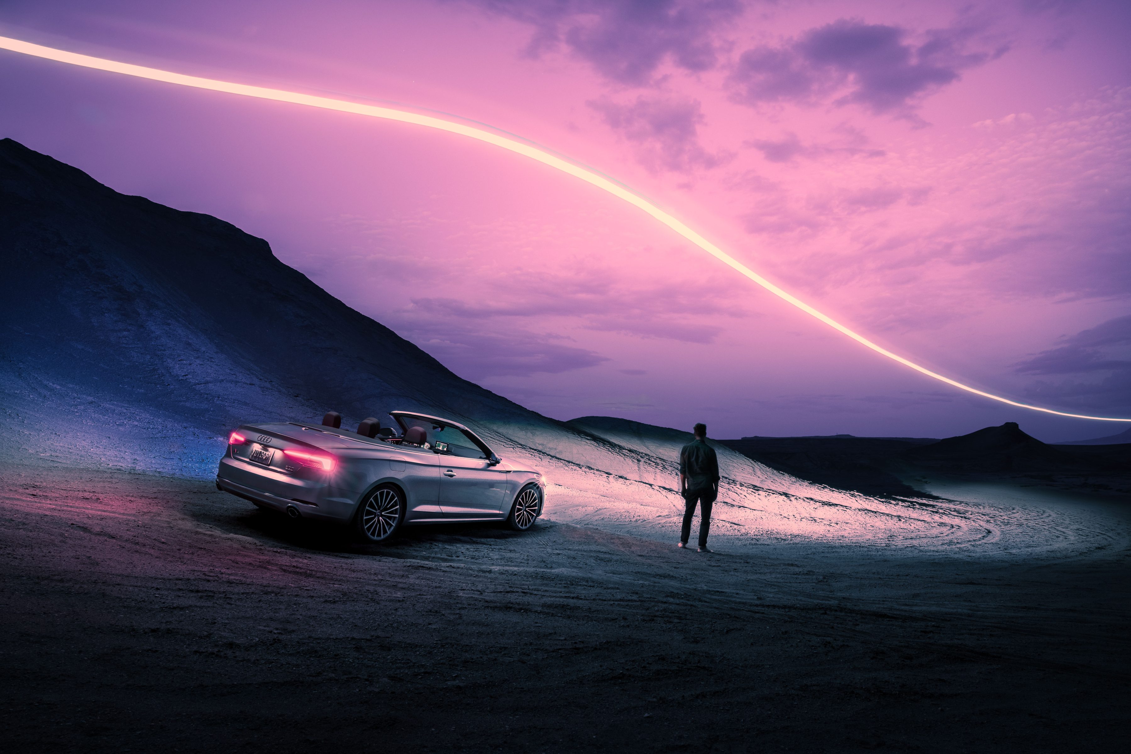 A photographer stands by a convertible car at dusk as a stunning drone light painting creates a bright trail across the purple sky.