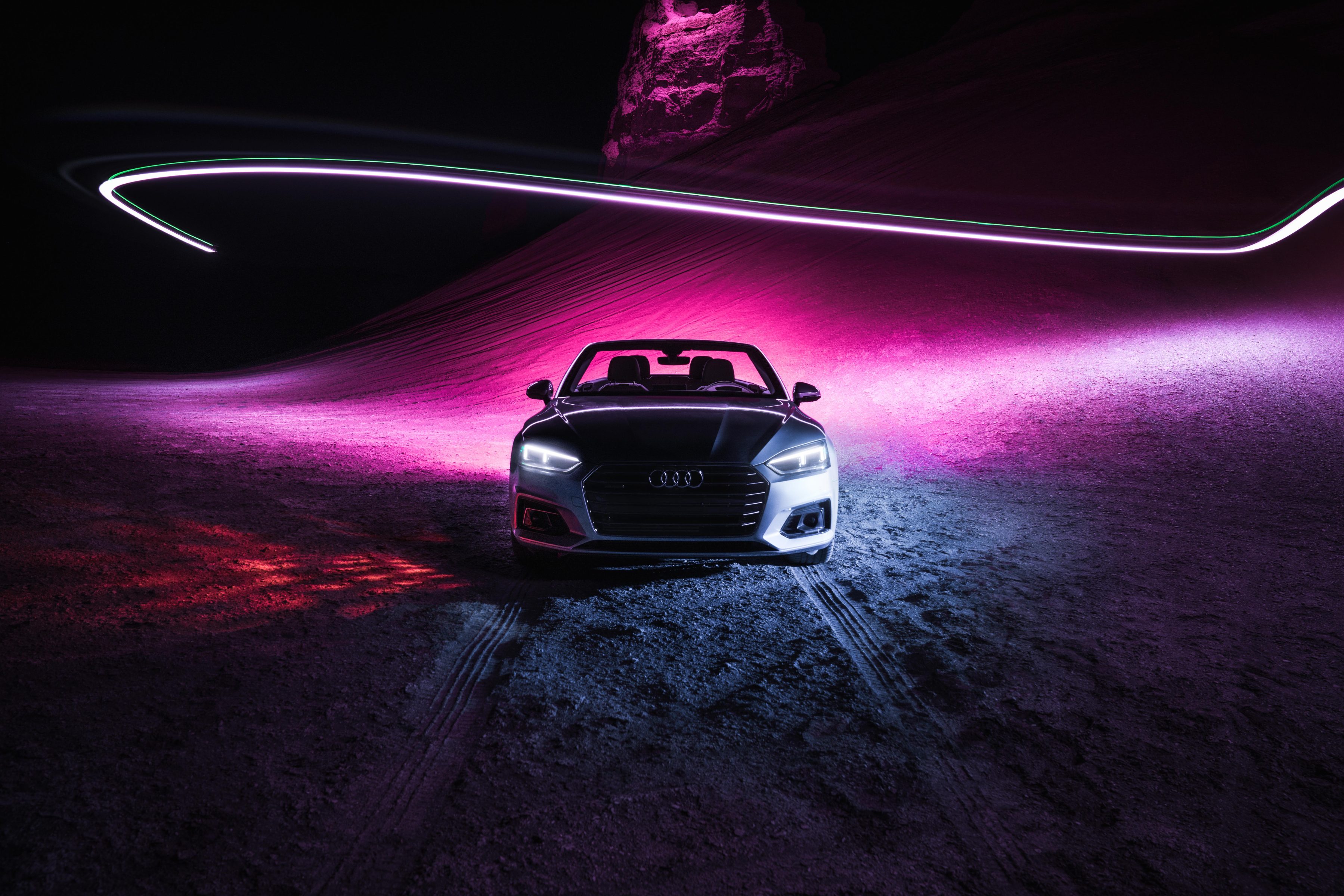 A futuristic Audi convertible car at night, illuminated by pink lights with dynamic drone light painting trails glowing in the background.