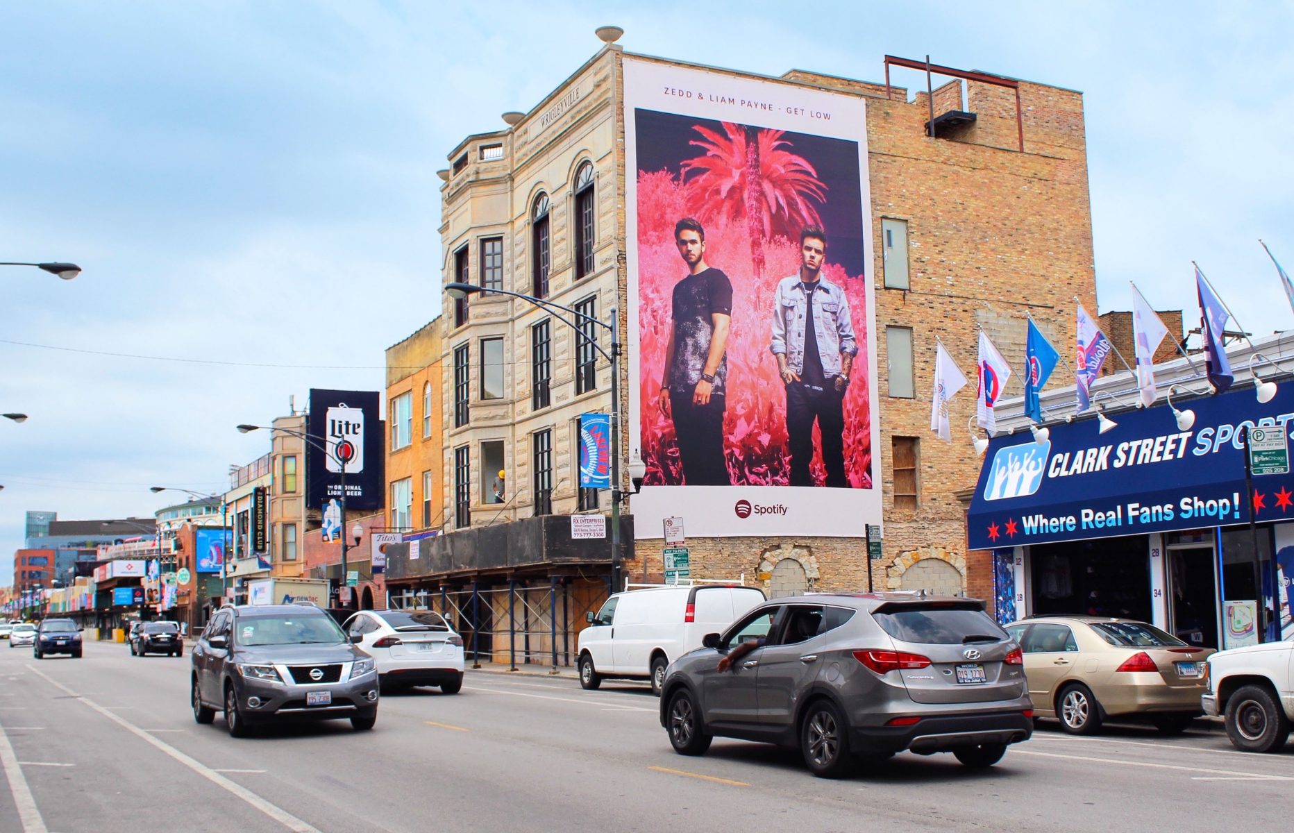 Cars driving down a city street with a large Spotify music billboard on a building, storefronts nearby, and a photographer capturing the vibrant urban scene.
