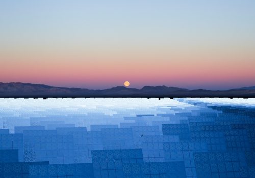 The full moon rises over distant mountains and a field of solar panels at sunset, as a photographer captures the scene enriched by mesmerizing drone light painting.
