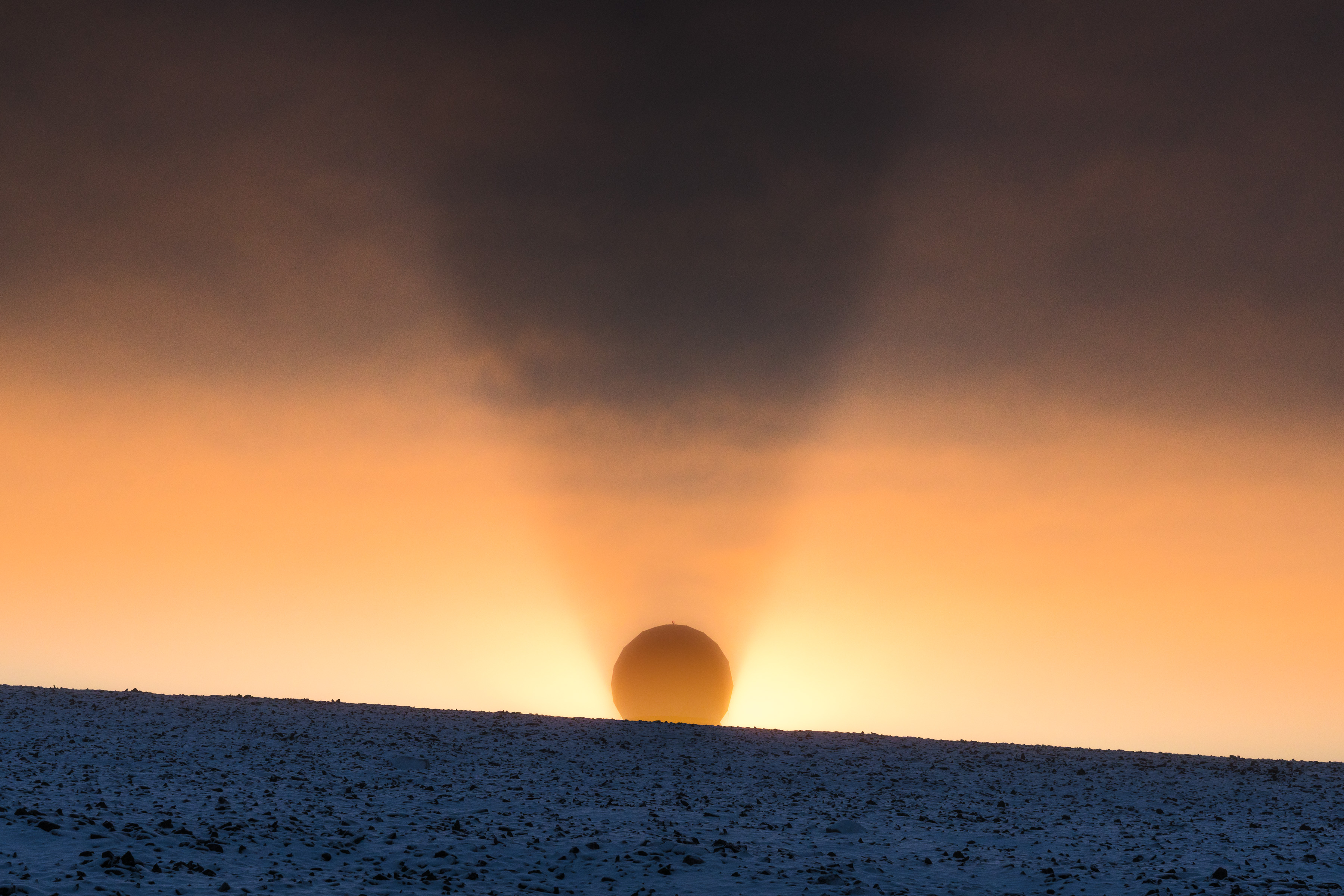 A round sun rises over a snowy hill, casting a dramatic shadow through the orange and blue sky, like a scene from drone light art.
