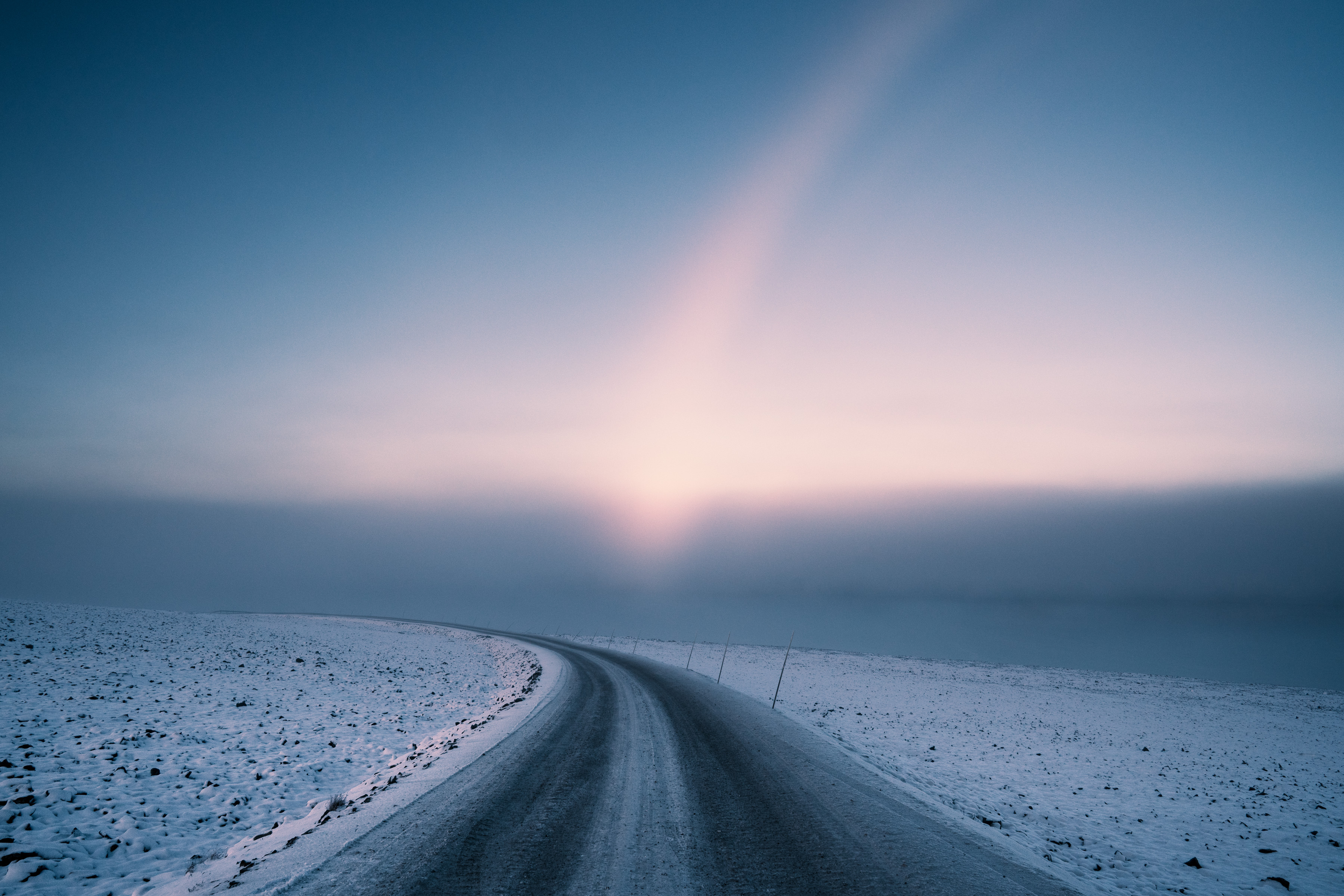 Snowy road curves through a winter landscape at dawn, with a pinkish halo around the rising sun, evoking the serene mood of fine artist nighttime landscape photography.