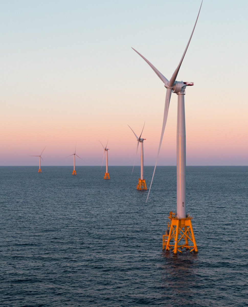 Five offshore wind turbines stand in the ocean at sunset, generating renewable energy, as a photographer captures their futuristic silhouette against the sky.