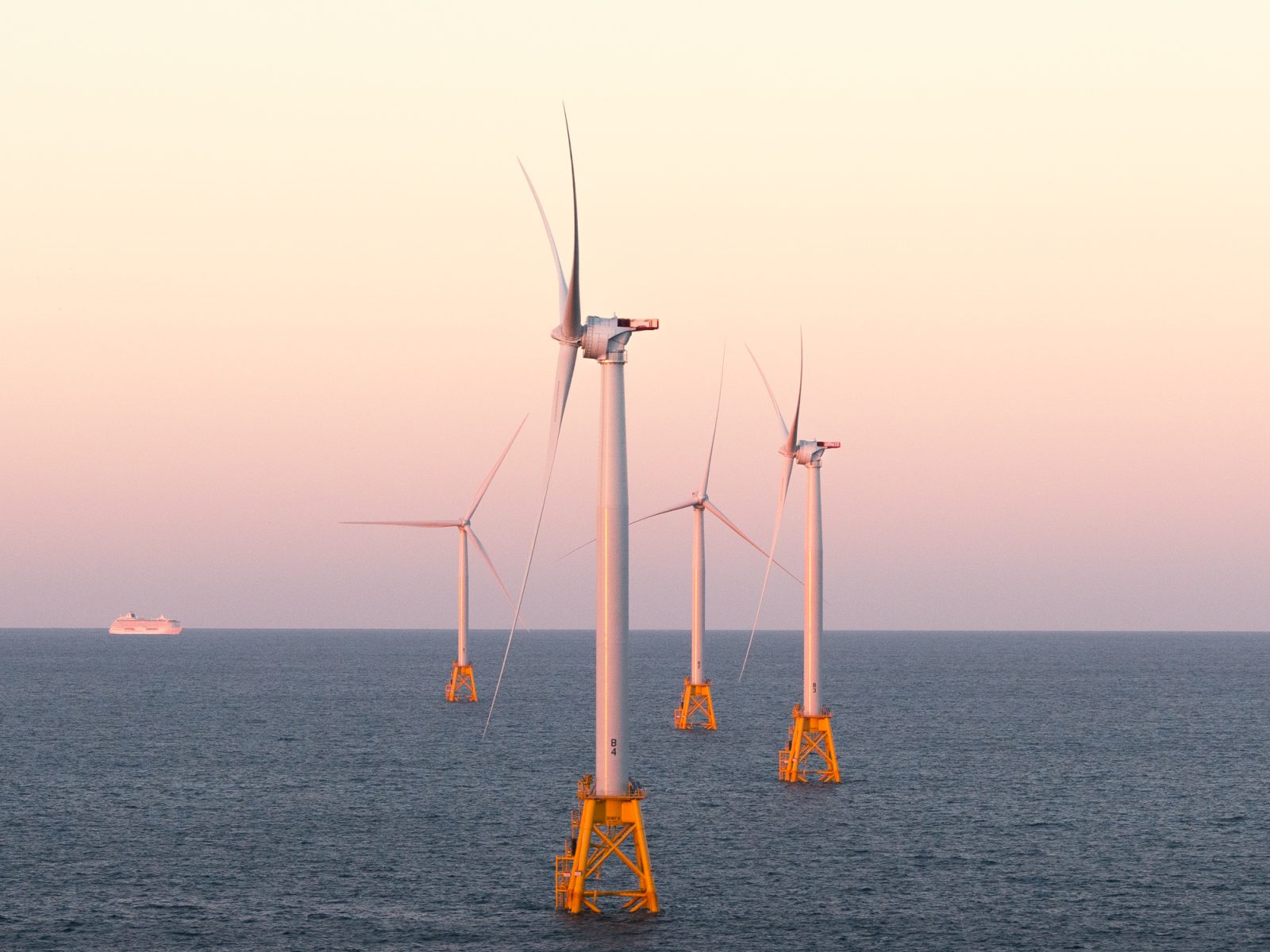 Four offshore wind turbines stand in the ocean at sunset, a distant cruise ship to the left. The scene feels futuristic, as if envisioned by a fine artist capturing tomorrow’s energy landscape.