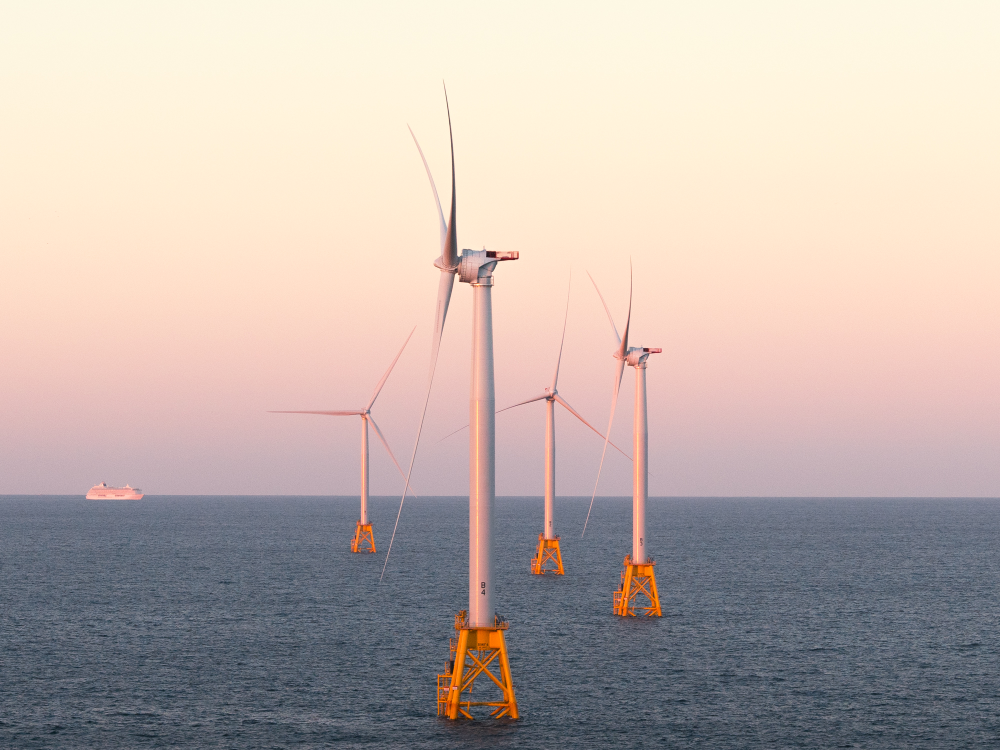 Four offshore wind turbines stand in the ocean at sunset, a distant cruise ship to the left. The scene feels futuristic, as if envisioned by a fine artist capturing tomorrow’s energy landscape.