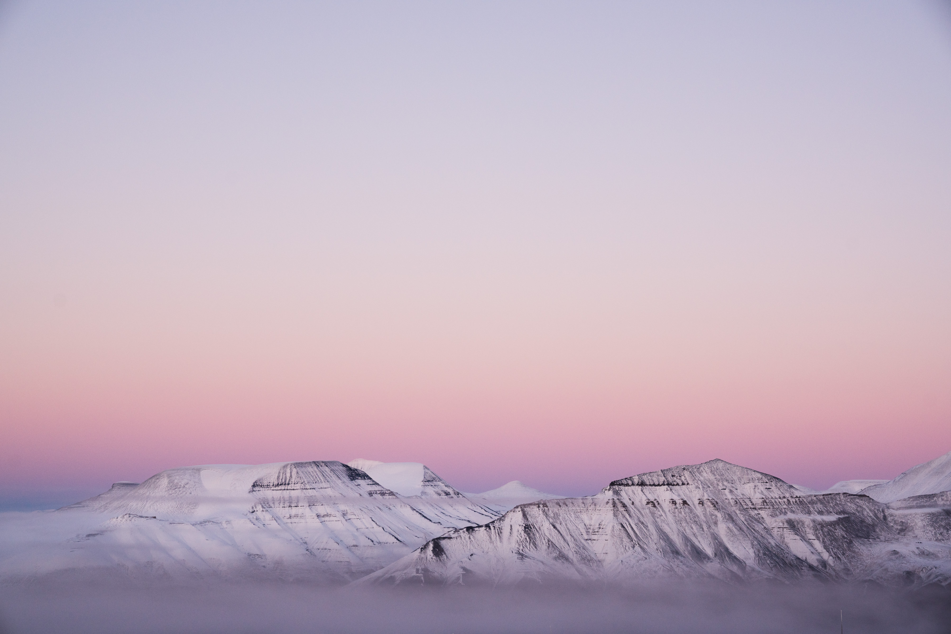 Snowy mountains under a pink and purple sunset sky, with mist at their base. A photographer captures the scene as futuristic drone light art dances above the peaks.
