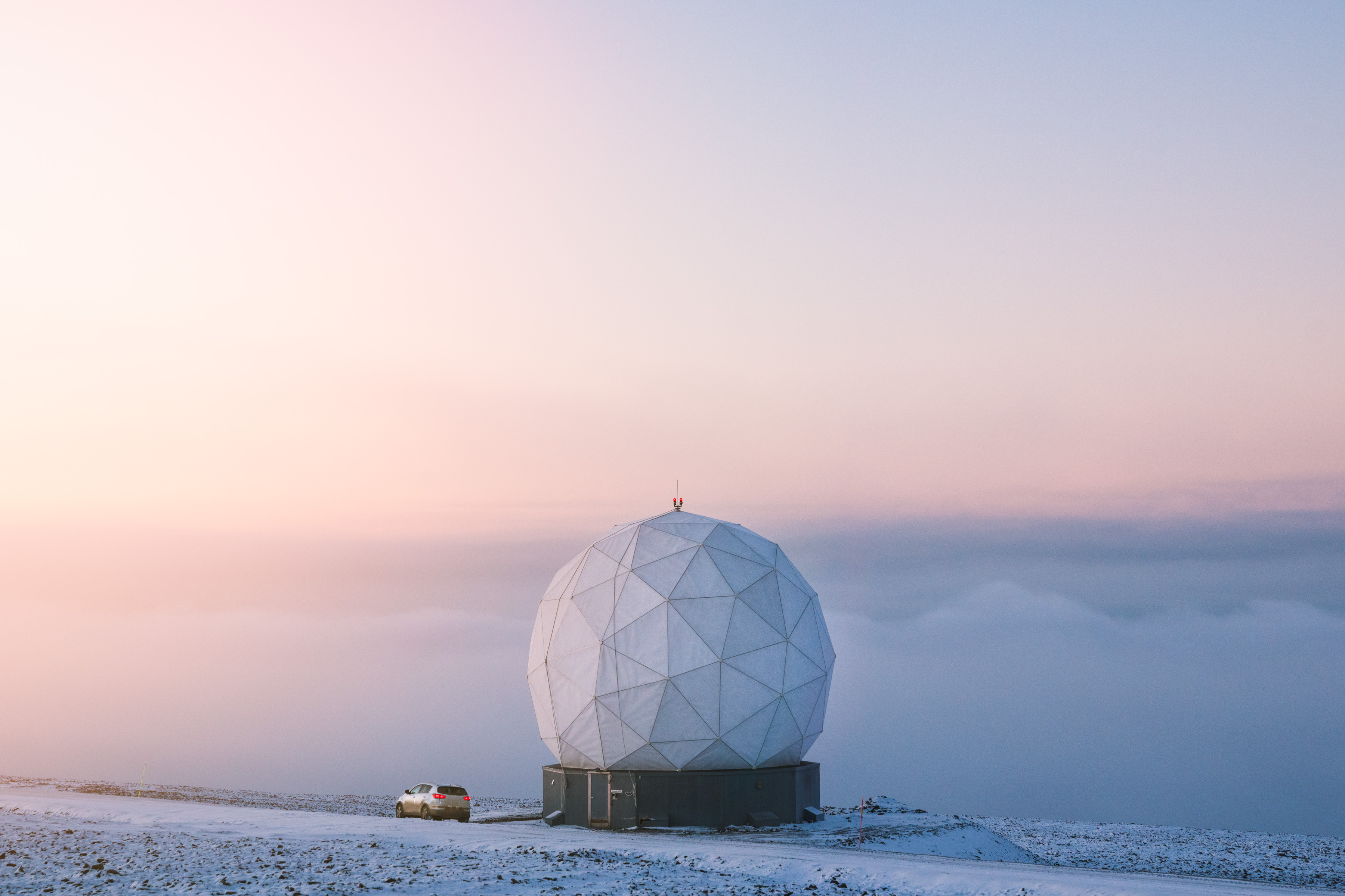 A white geodesic dome and a car sit on a snowy landscape, illuminated by the gentle hues of sunrise or sunset—an inspiring scene for any photographer who appreciates ethereal light and dramatic compositions.