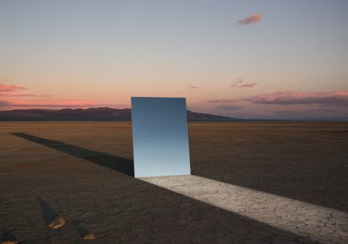 A tall mirror stands on cracked desert ground, reflecting a starry sky, with a long shadow at sunset—a photographer captures the scene, hinting at a futuristic world.