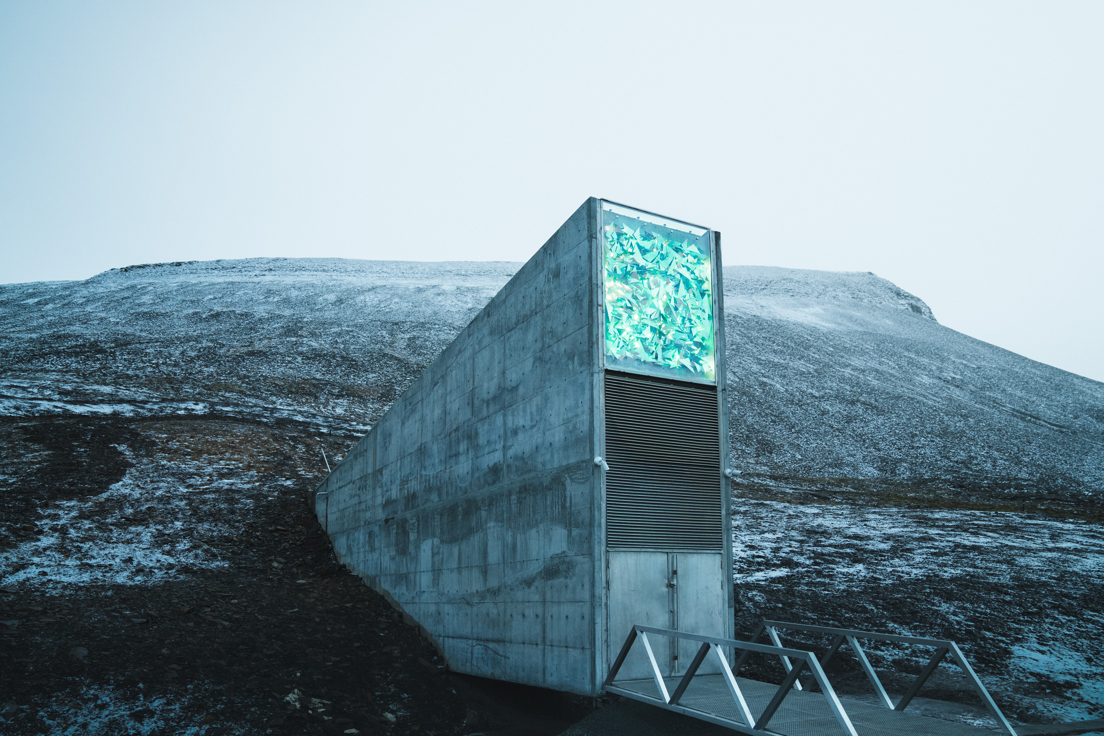 A futuristic concrete structure set into a snowy hillside under a pale, overcast sky, illuminated by ethereal patterns of drone light art.