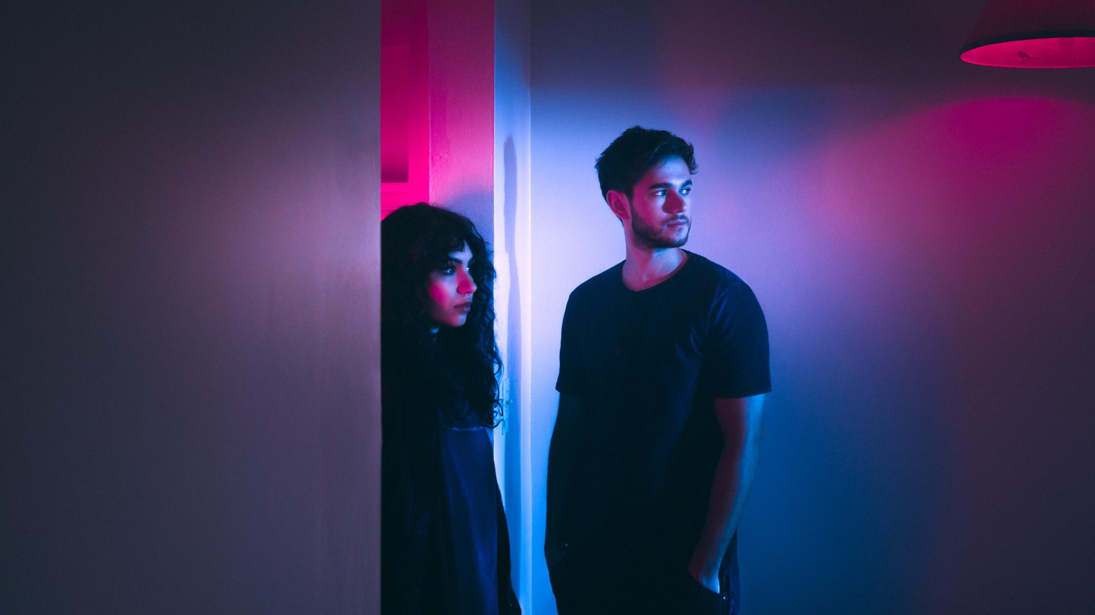 Two people stand in a dimly lit room with blue and pink neon lighting, evoking a futuristic vibe reminiscent of drone light art displays. They look in different directions, immersed in the ambient glow.