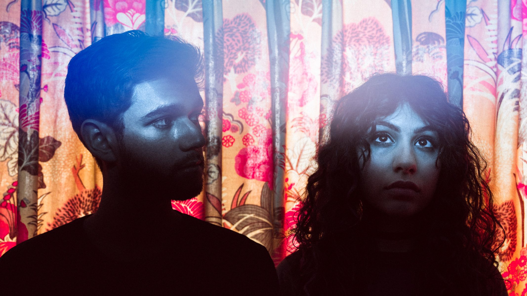 Two people stand in dramatic lighting against a colorful floral curtain, evoking the atmosphere of nighttime landscape photography as they look in different directions.