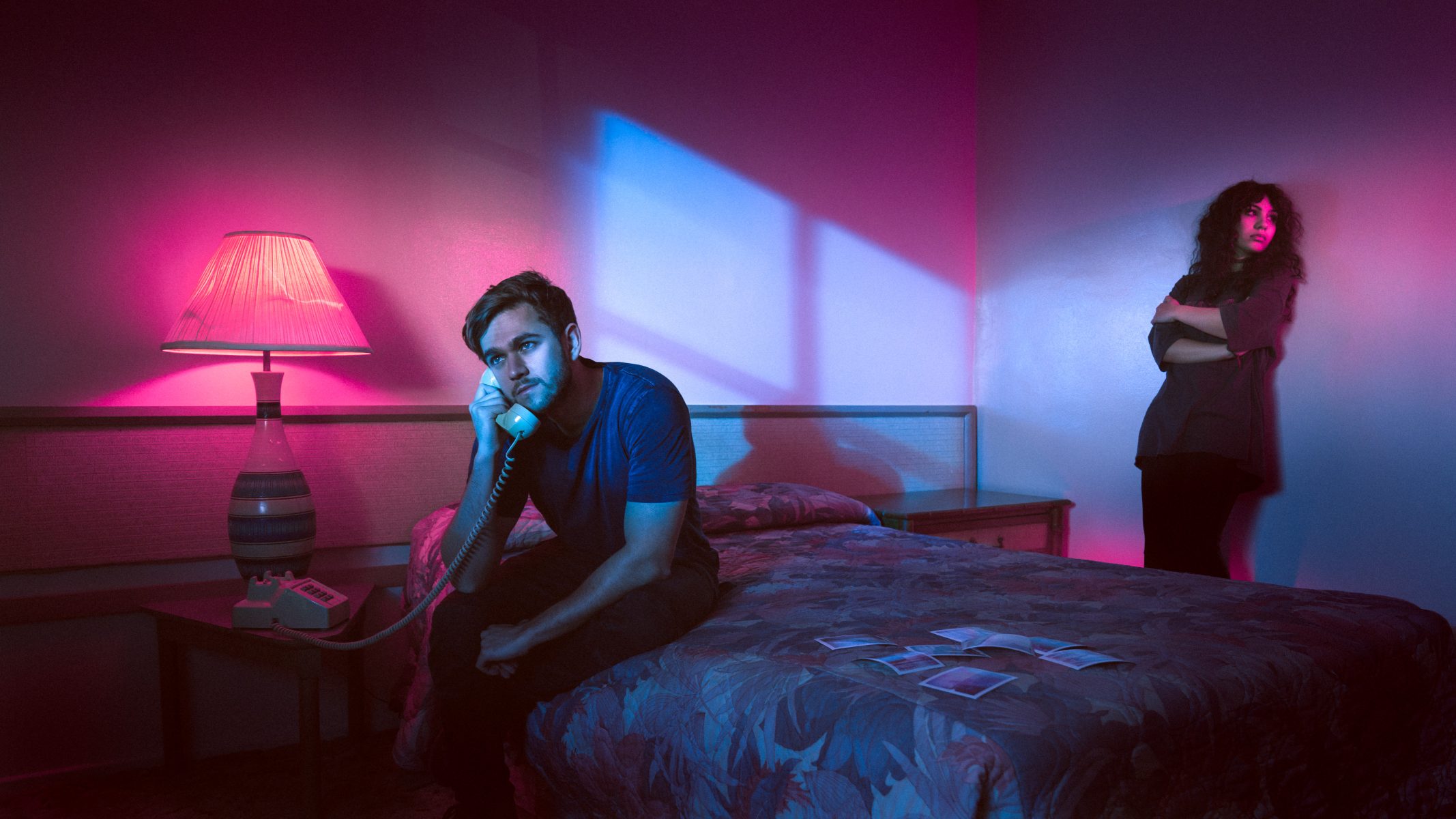A man sits on a bed talking on the phone, while a woman stands in the background of a neon-lit, futuristic room—captured by a photographer with vibrant drone light painting effects illuminating the scene.