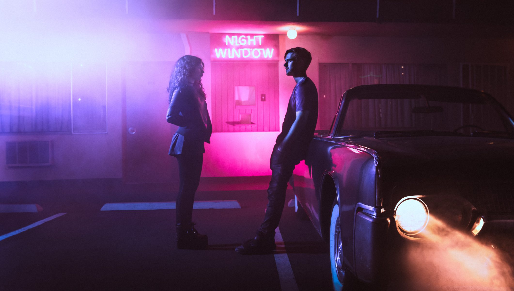 Two people stand by a car at night, neon Night Window sign glowing behind them, surrounded by mist—a futuristic scene reminiscent of nighttime landscape photography.