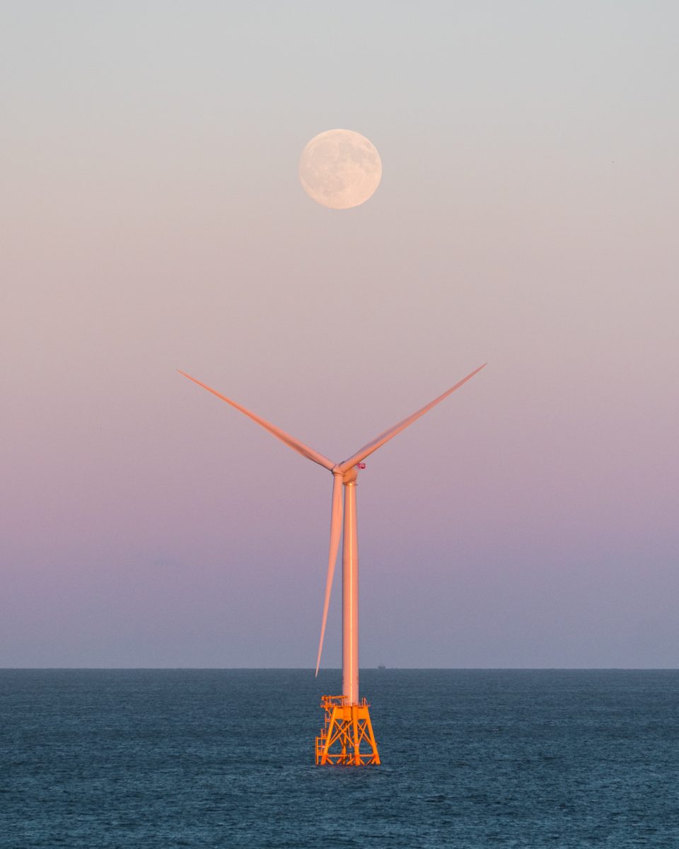 A wind turbine at sea stands beneath the full moon rising in a pastel sky, illuminated by futuristic drone light painting that dances above the waves.
