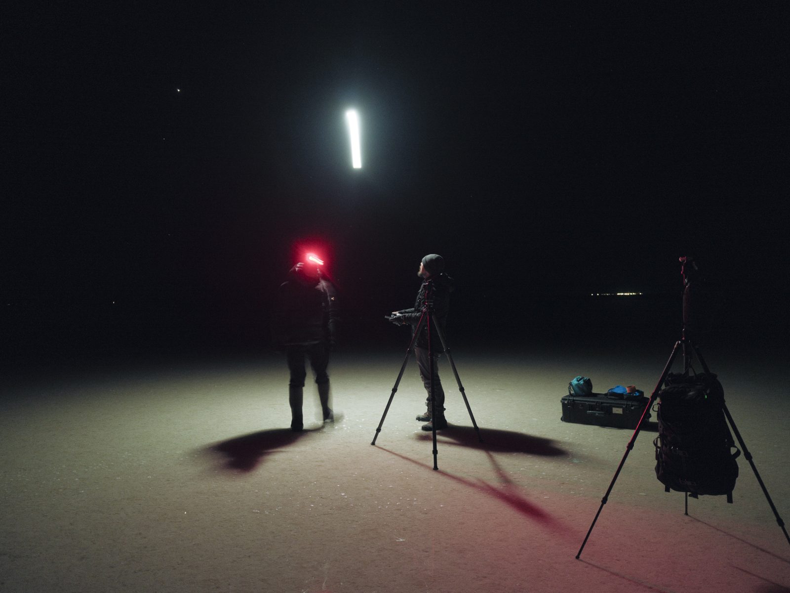 Two people, perhaps a photographer and a fine artist, stand in the dark with tripods and gear, illuminated by a bright light in the night sky—a perfect scene for nighttime landscape photography.