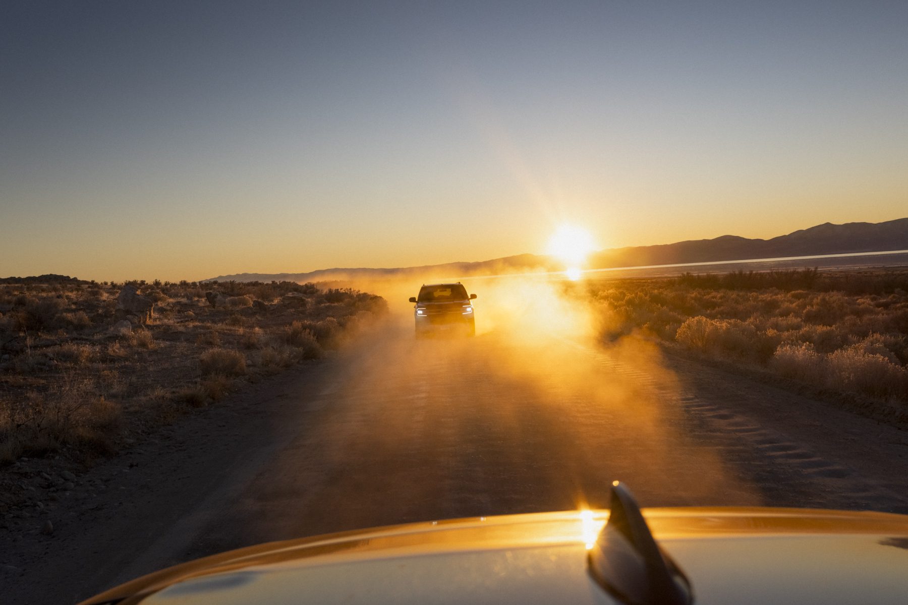 A car drives down a dusty road at sunset, headlights on, while distant mountains glow under the fading sun like a scene from drone light painting.