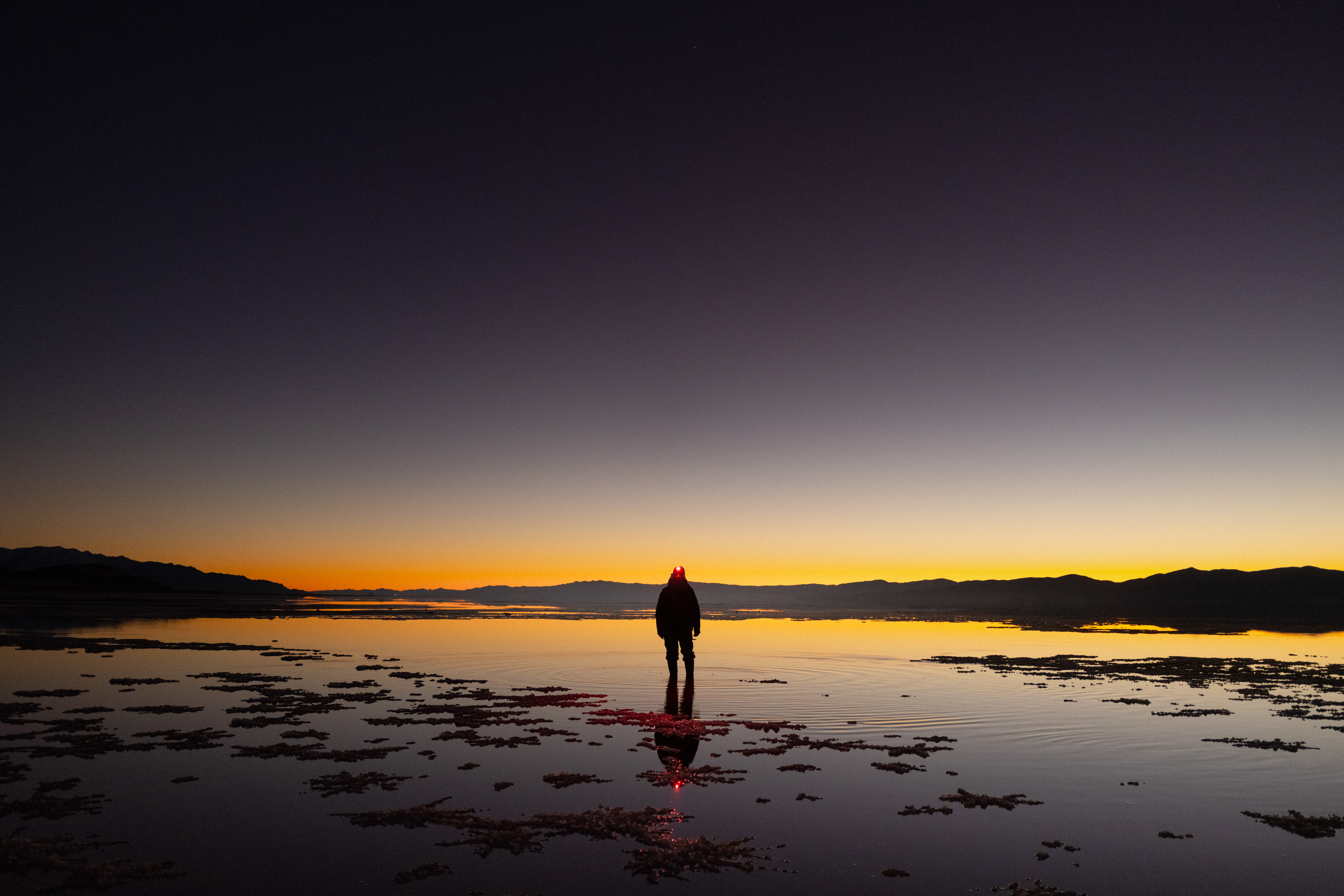 Silhouette of a person standing on a reflective lake at sunset with mountains in the background, as a photographer captures the breathtaking scene.
