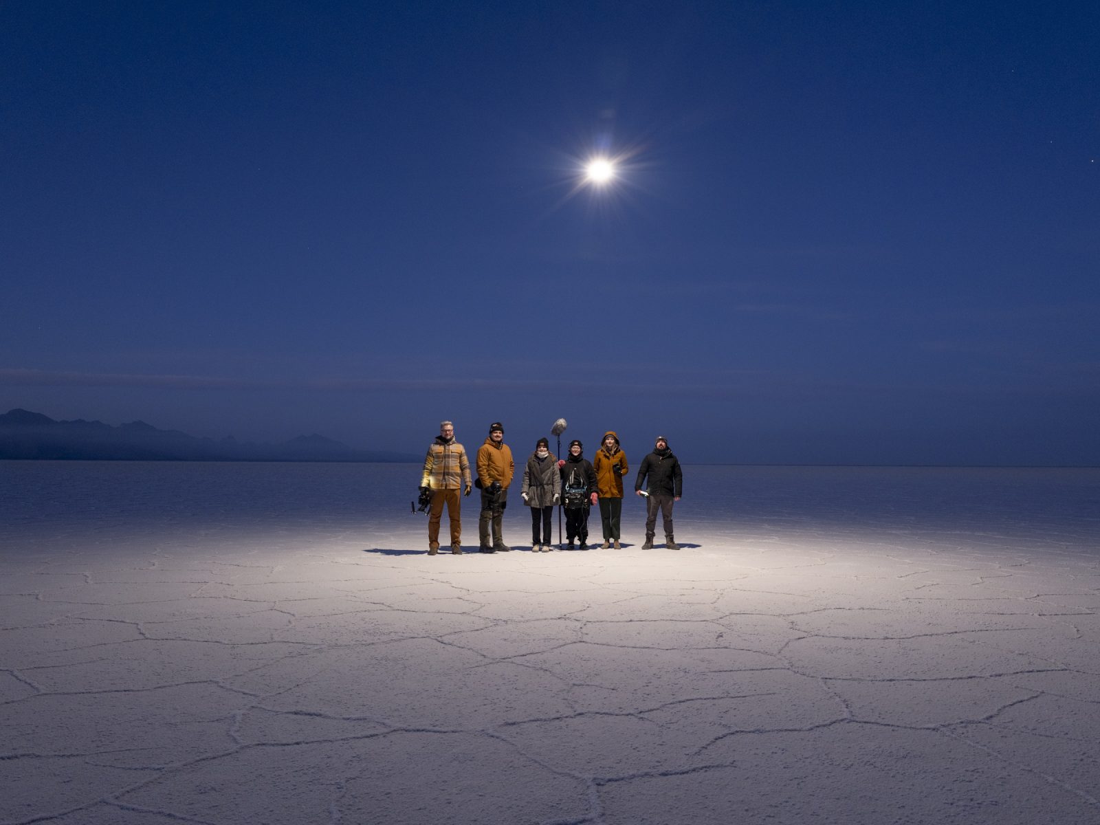 Seven people stand together on a cracked salt flat at night under a bright full moon, capturing the beauty of nighttime landscape photography.