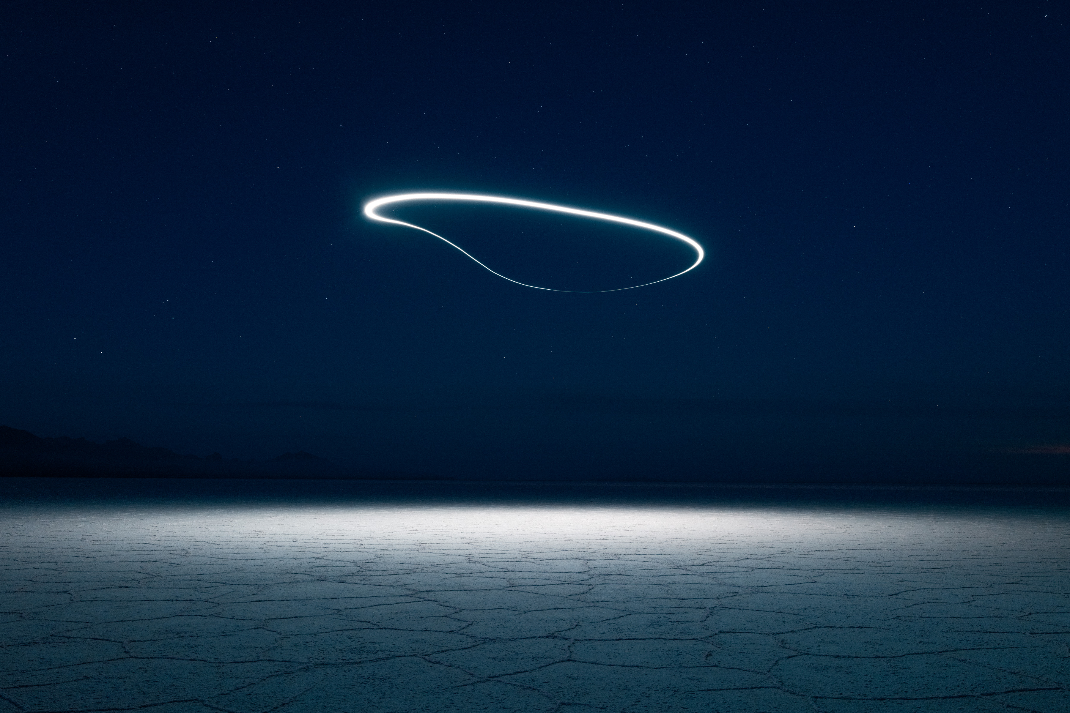 A glowing loop of light, created by drone light painting, hovers in the night sky above a cracked, dry landscape.