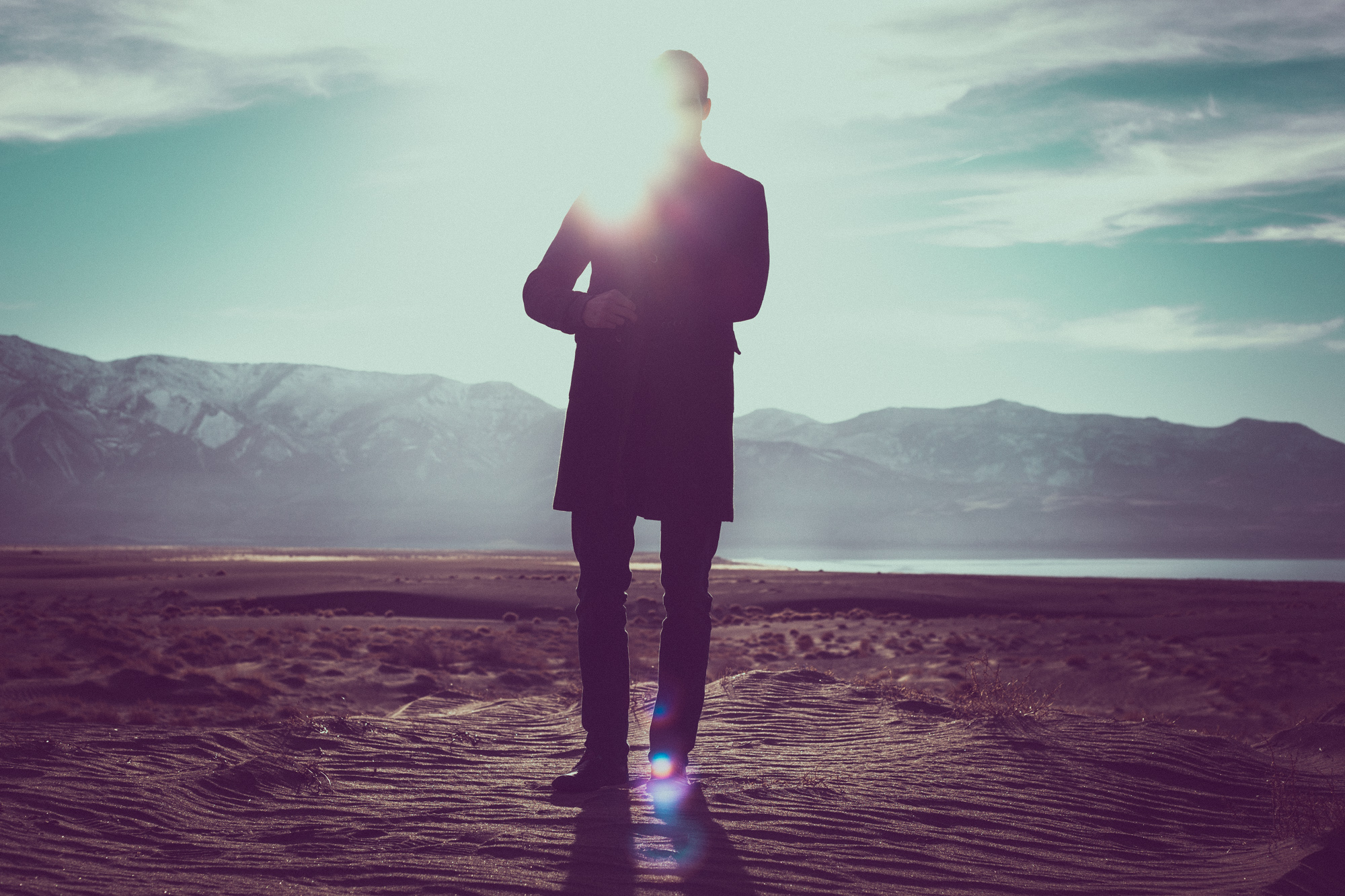 Silhouette of a person standing on sand with mountains and sunlight in the background, capturing the essence of drone light painting for a mesmerizing nighttime landscape photography scene.