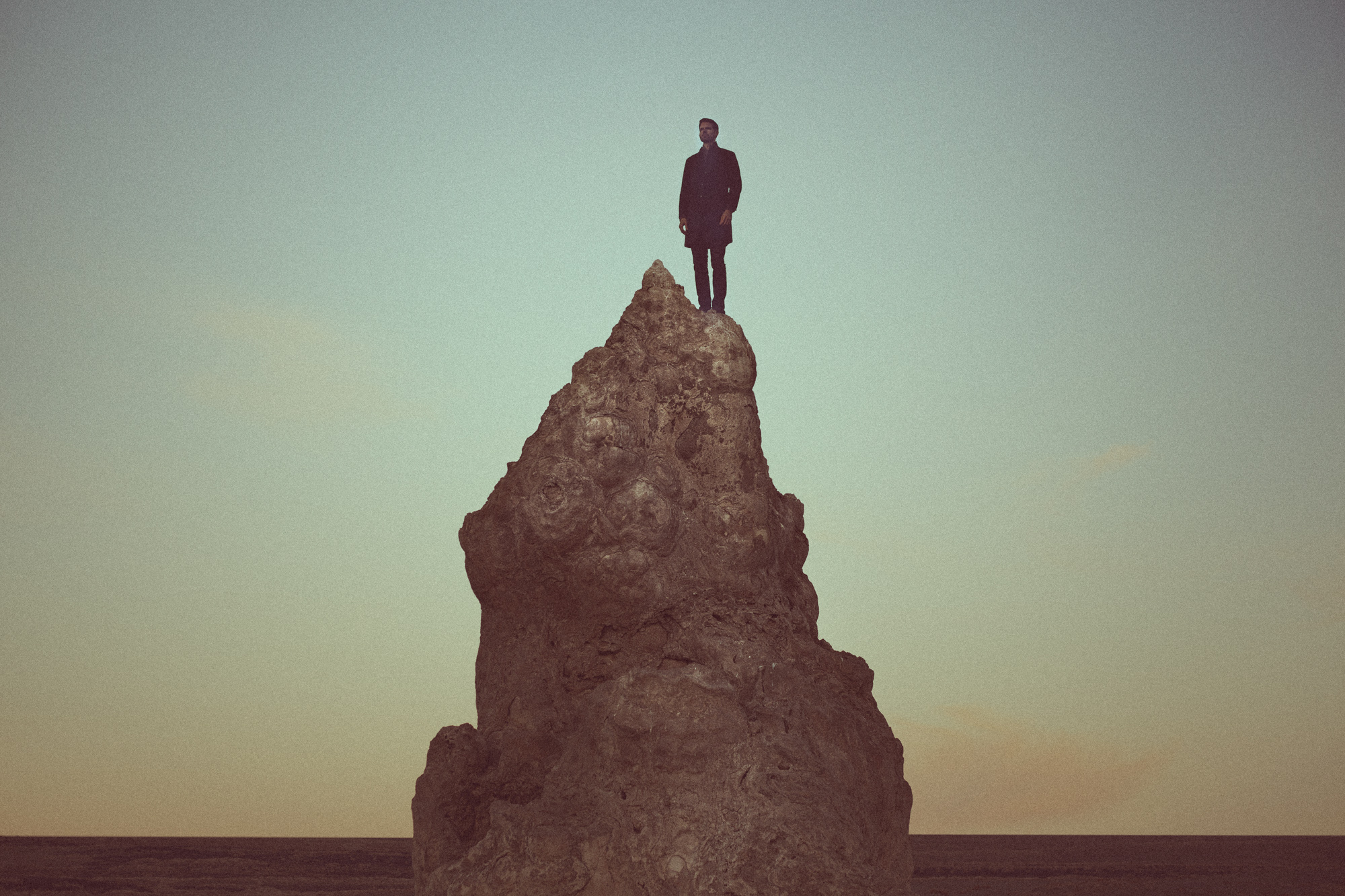 A photographer stands on top of a tall, jagged rock formation under a pale sky at sunset, capturing the futuristic glow of drone light painting swirling above.