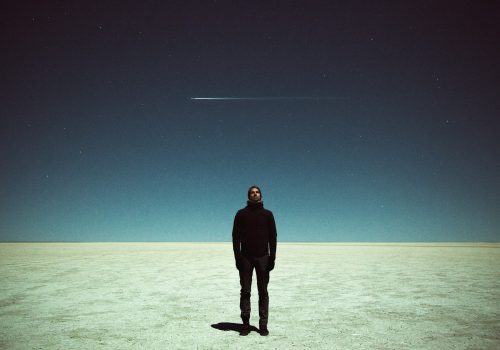 A photographer stands alone on a vast, barren plain under a starry sky, capturing the moment as a shooting star streaks above.