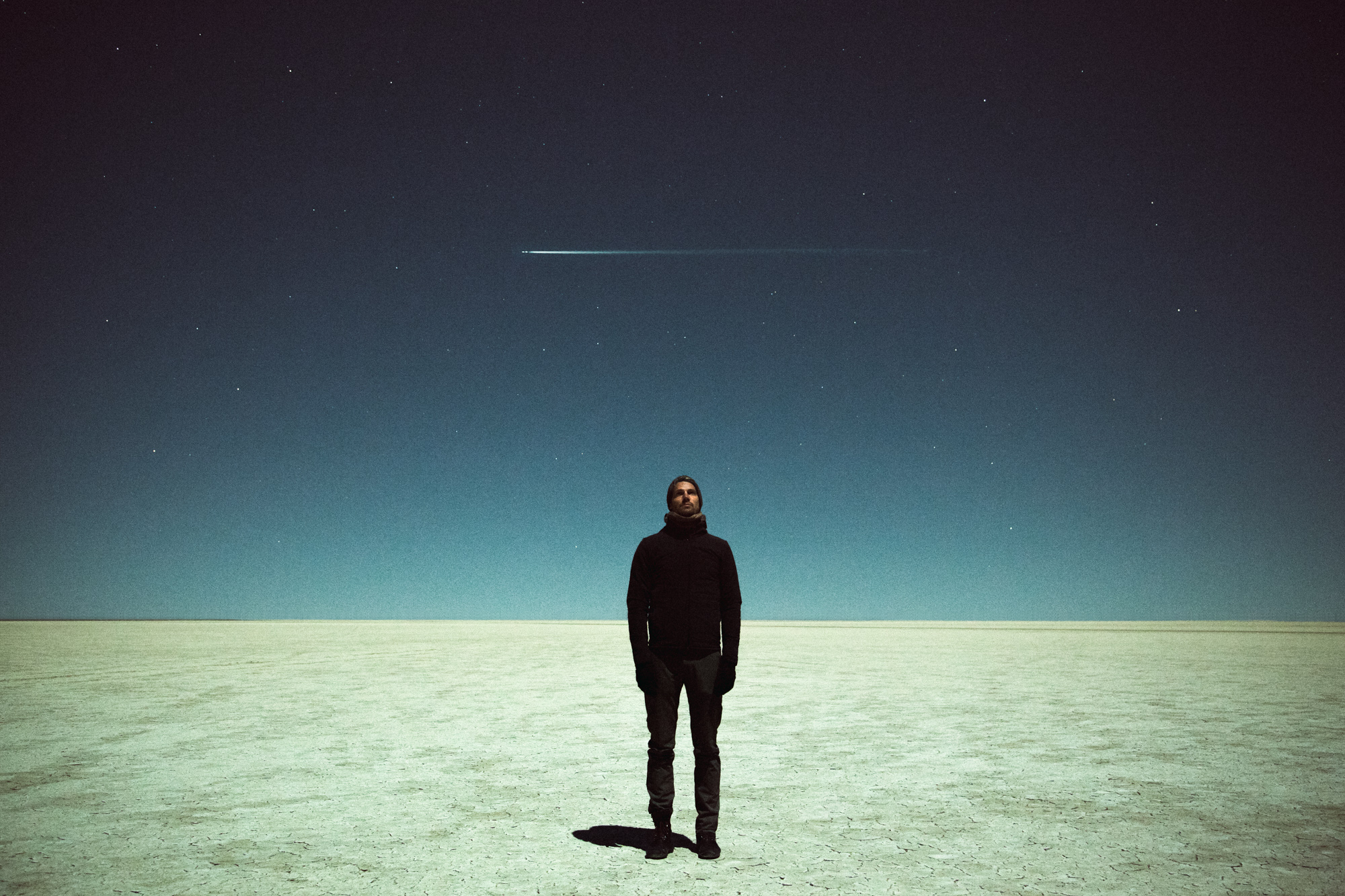 A photographer stands alone on a vast, barren plain under a starry sky, capturing the moment as a shooting star streaks above.