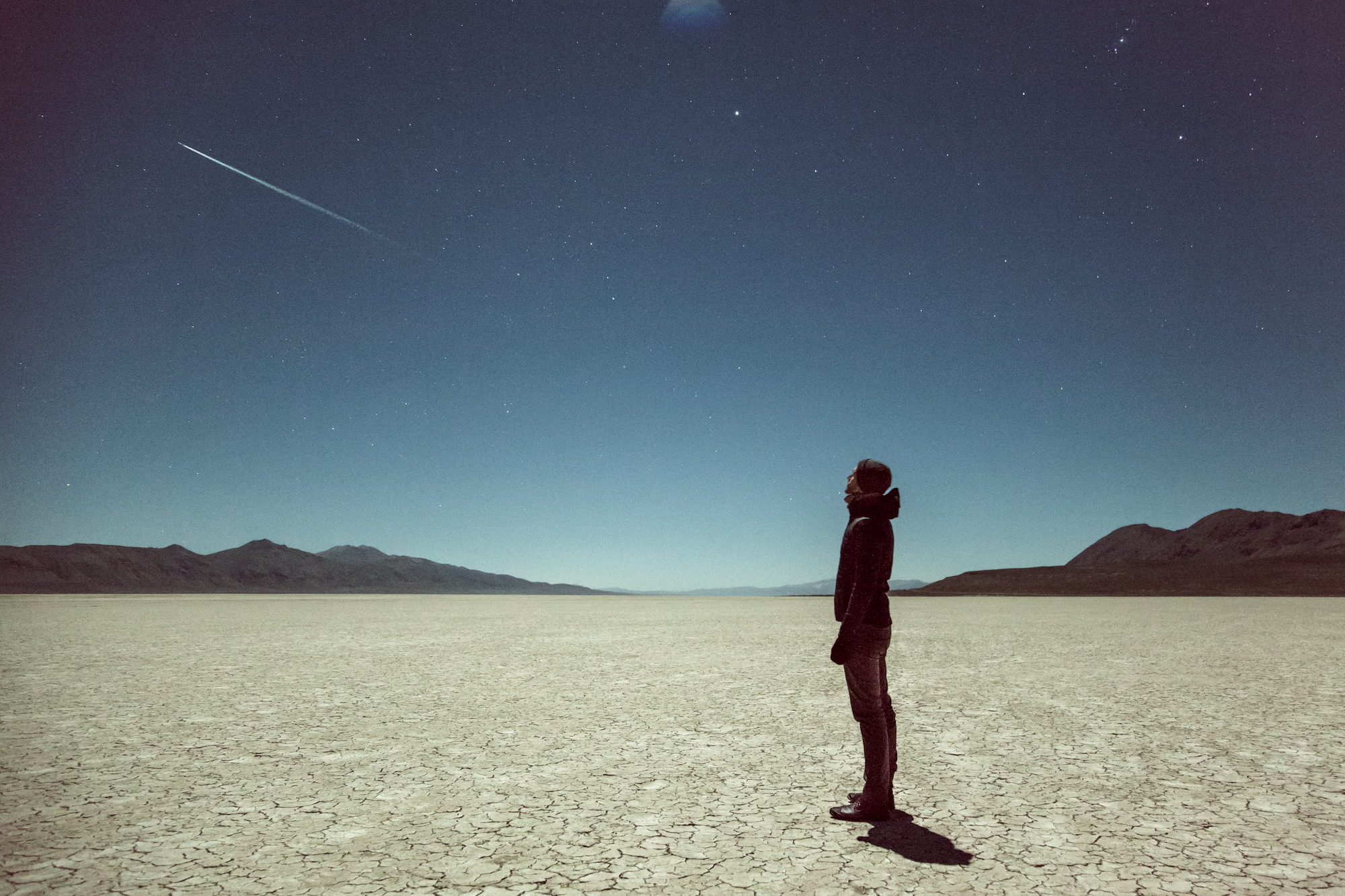 A person stands on cracked desert ground at night, gazing up at a shooting star in a clear, starry sky—a scene reminiscent of nighttime landscape photography by a fine artist.