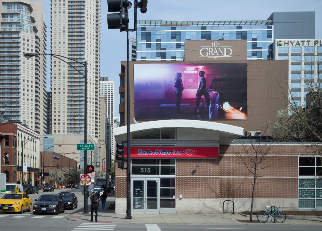 A city street with a Bank of America and a large digital billboard above displaying people walking, illuminated in a style reminiscent of nighttime landscape photography.