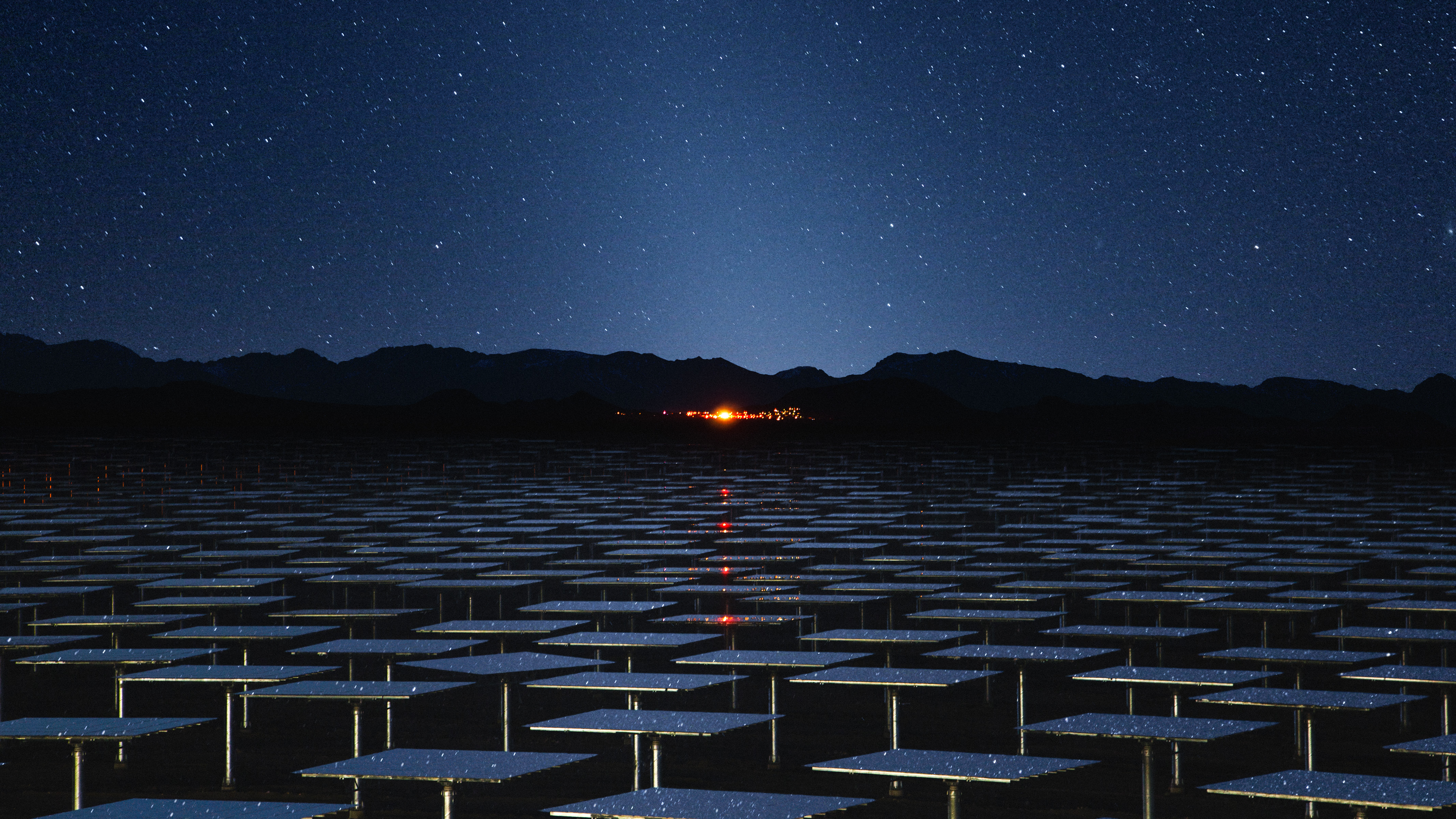 Solar panels under a starry night sky with distant mountains and city lights glowing on the horizon, captured with the eye of a fine artist specializing in nighttime landscape photography.