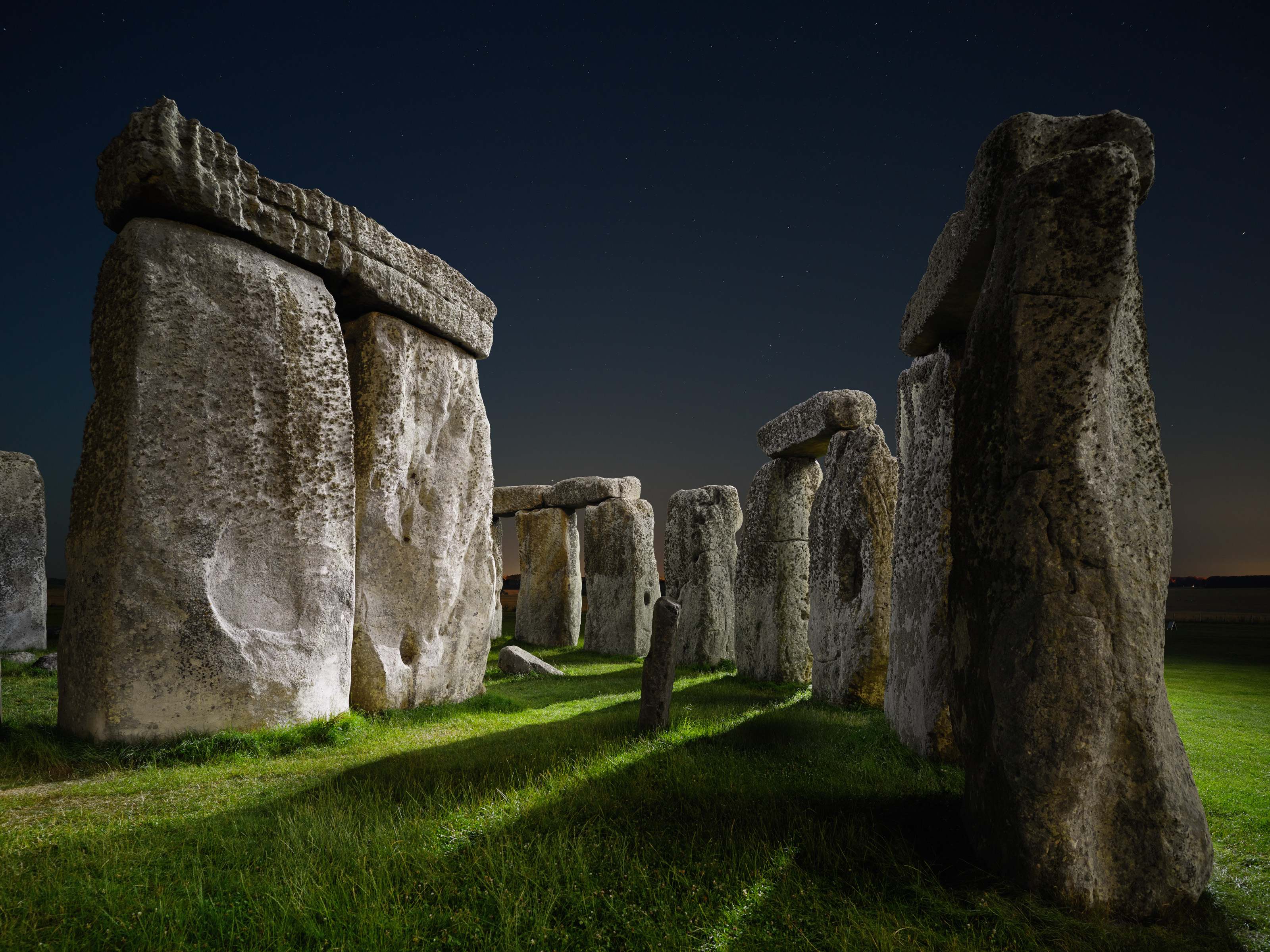Stonehenge standing stones at night, illuminated by soft lighting in a striking example of nighttime landscape photography, casting long shadows on the grass.