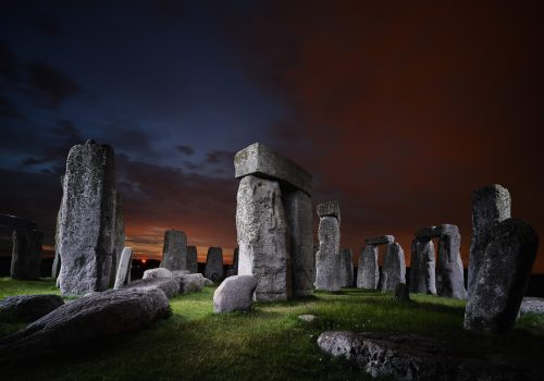 Large stone structures of Stonehenge at dusk, illuminated by drone light art against a dramatic dark sky and vivid orange clouds in the background.