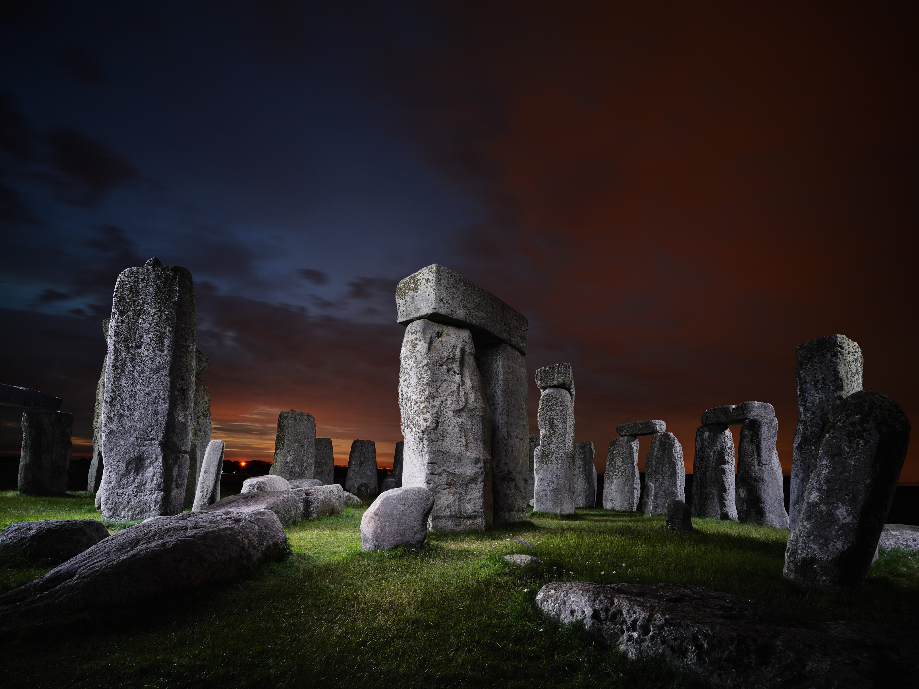 Large stone structures of Stonehenge at dusk, illuminated by drone light art against a dramatic dark sky and vivid orange clouds in the background.