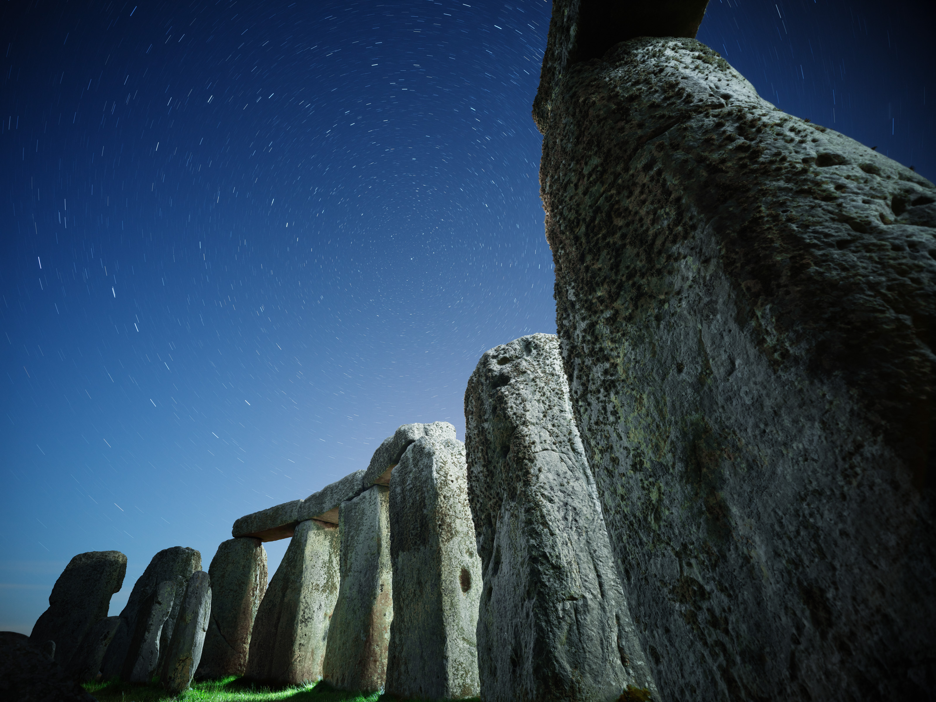 Large ancient stone pillars beneath a starry night sky, where circular star trails blend seamlessly with mesmerizing drone light art, creating a surreal nighttime landscape photography scene.
