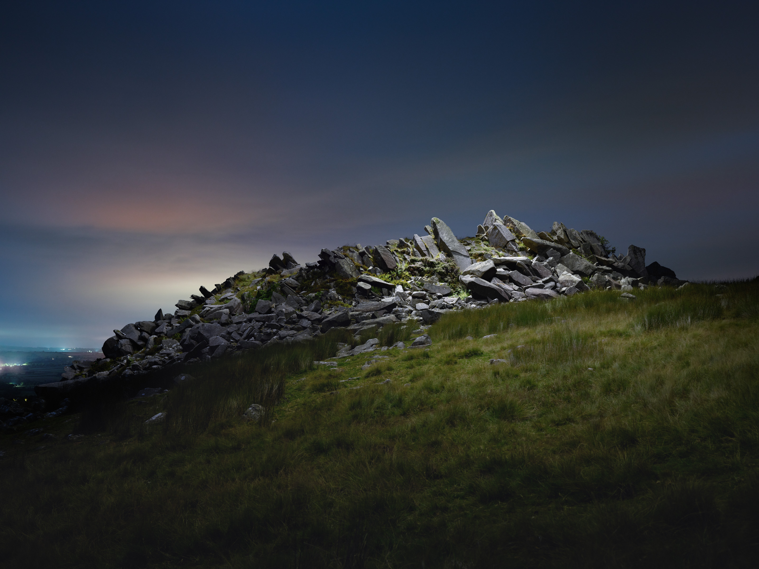 A rocky hillside illuminated at night with grass in the foreground and a soft, glowing sky in the background creates a futuristic scene, perfect for a photographer capturing mesmerizing drone light art.