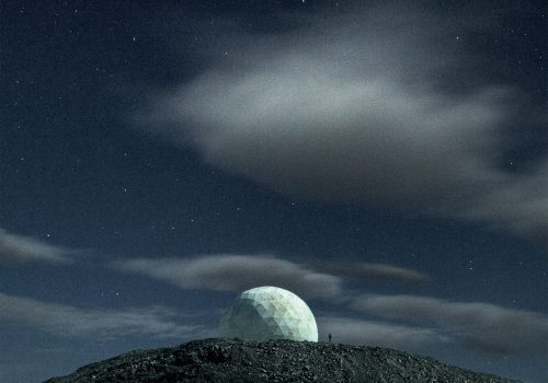 A geodesic dome on a hilltop under a starry night sky with a few scattered clouds, captured by a photographer as vibrant drone light art illuminates the scene.