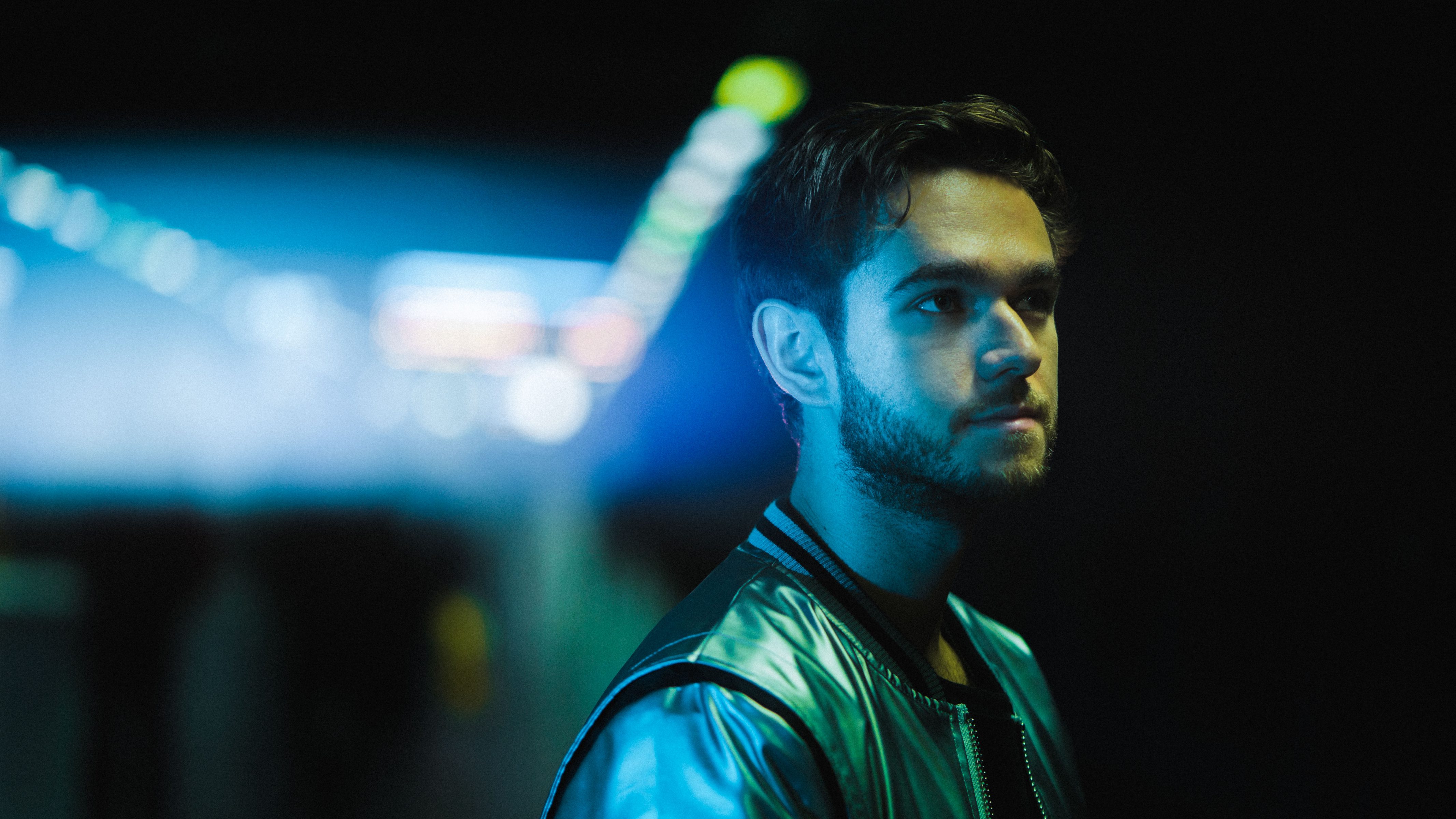 Young man with a beard in a shiny jacket stands in a dimly lit area with blurred blue and yellow lights, evoking the vibrant glow of drone light painting.