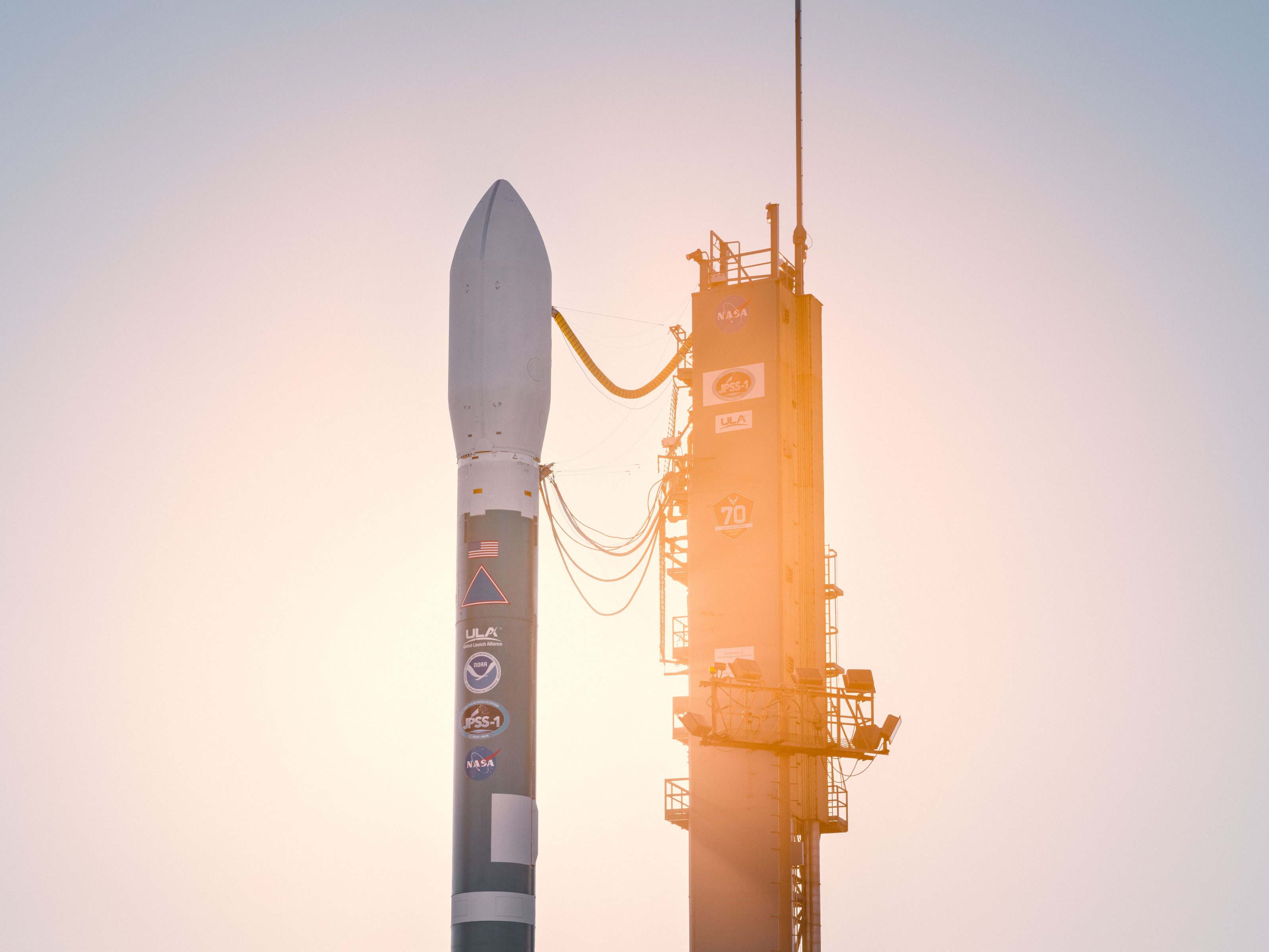 A rocket stands on its launch pad at sunrise, bathed in warm light, with logos visible on its side as a photographer captures the scene, complemented by striking drone light art in the sky above.