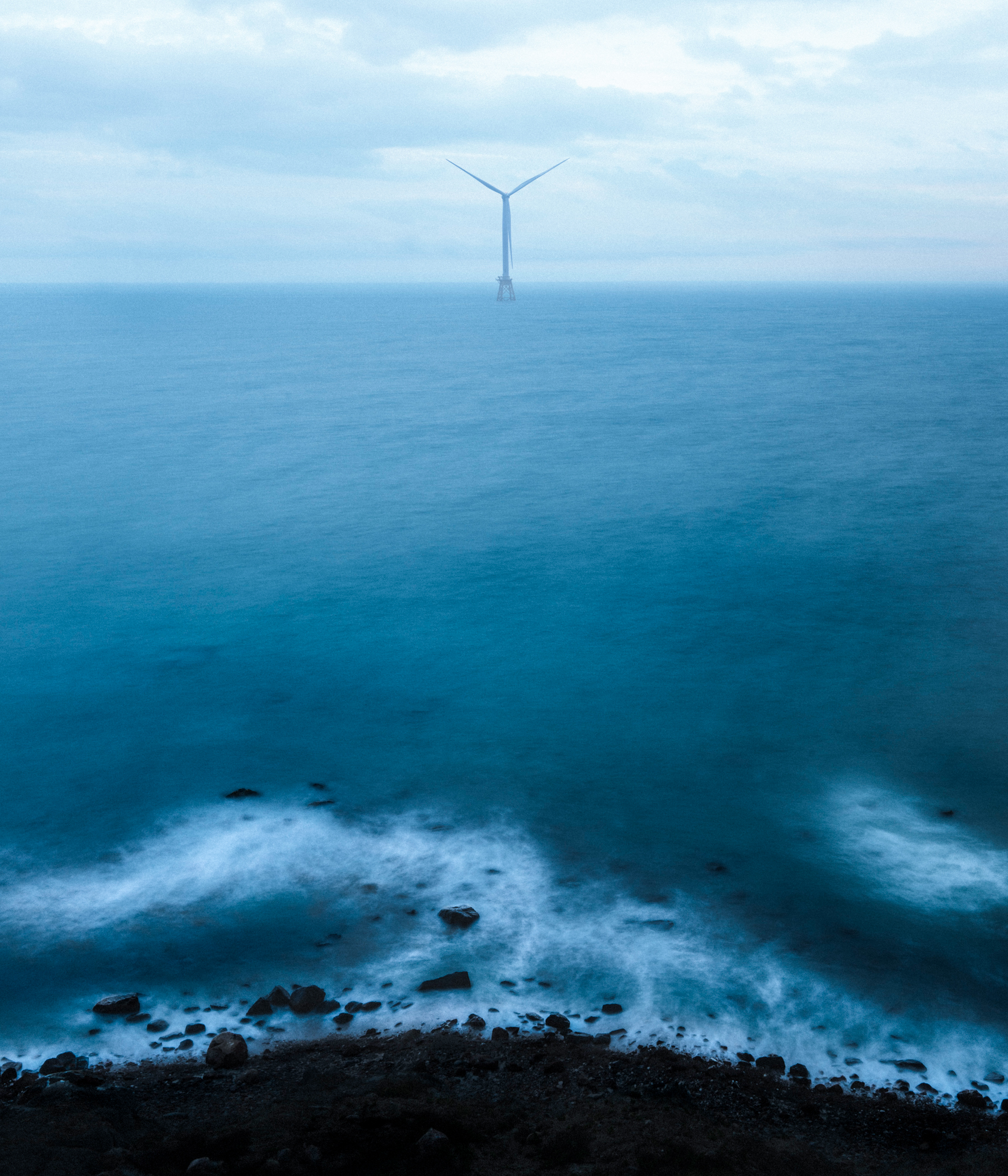 A lone wind turbine stands in the ocean beneath a cloudy sky, illuminated by drone light art, with waves crashing on a rocky shore—a striking scene of futuristic nighttime landscape photography.