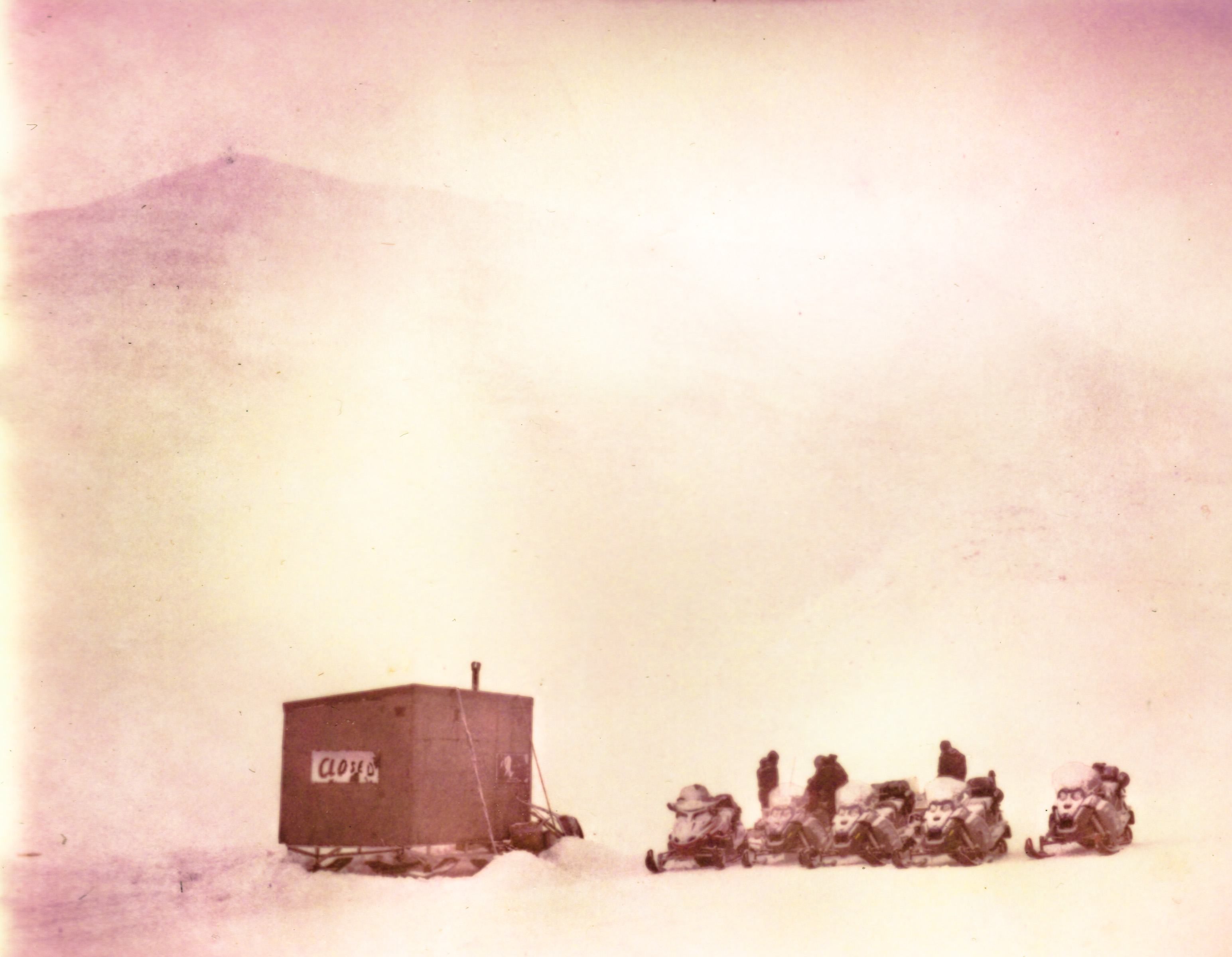 A small group on snowmobiles beside a hut in a snowy, foggy landscape, with swirling patterns of drone light painting illuminating the winter night.