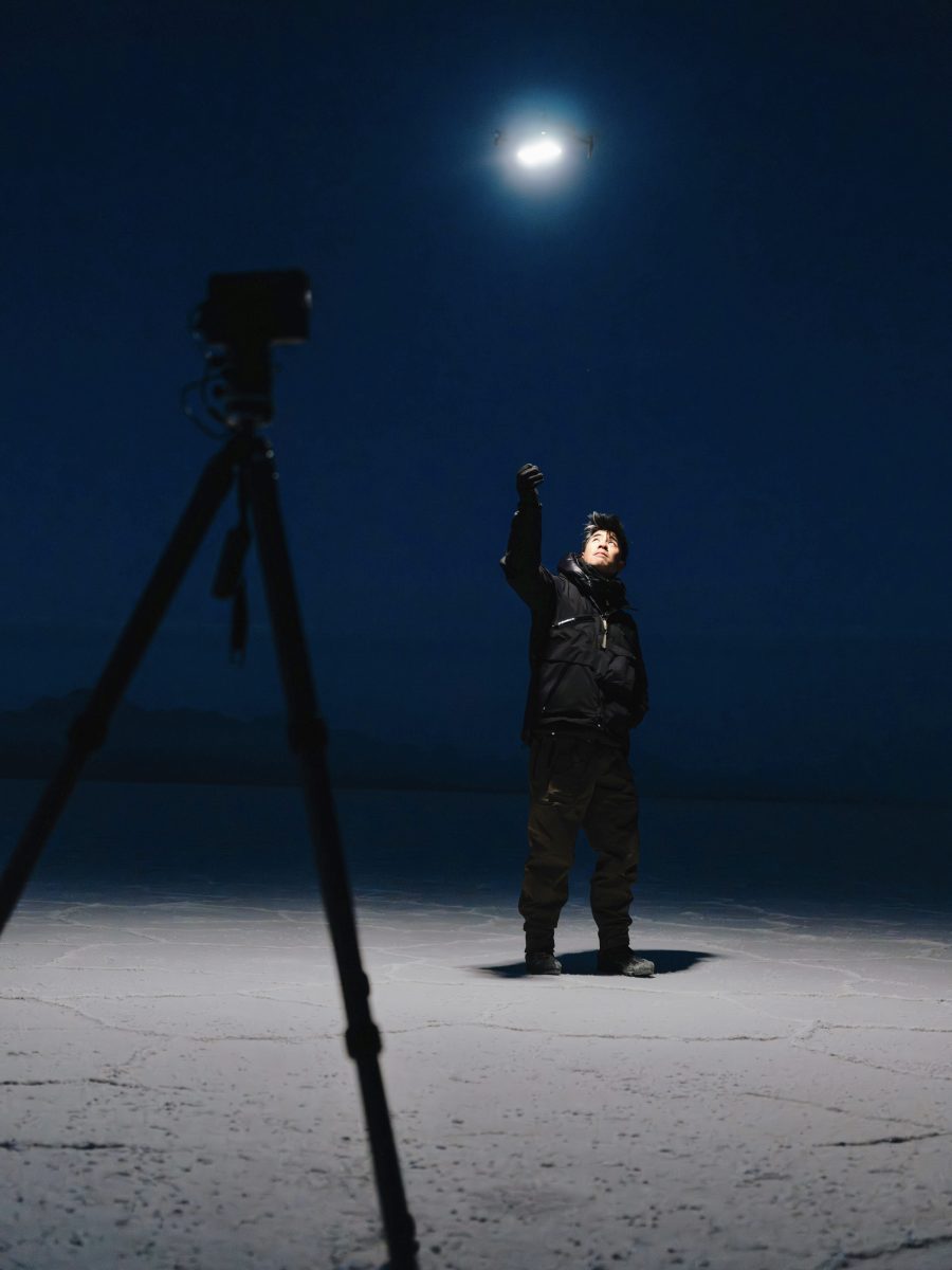 A photographer stands on a flat, cracked surface at night, reaching toward a bright drone light in a scene that feels futuristic; tripod in foreground.