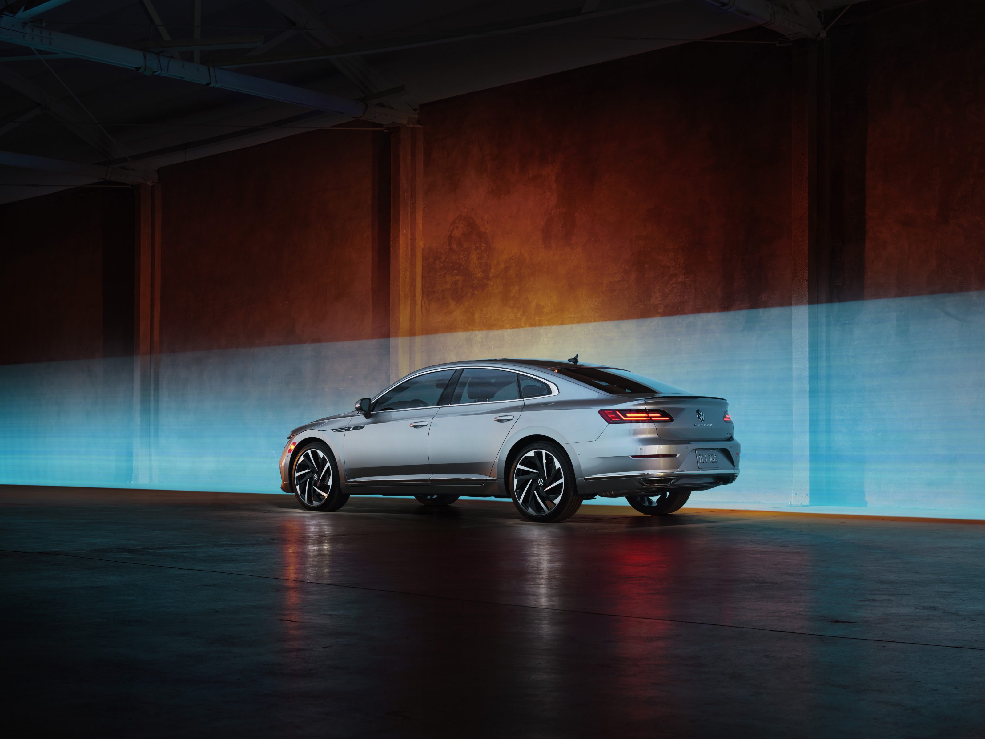 Silver sedan with black rims parked indoors against a blue and orange gradient-lit wall, reminiscent of drone light art crafted by a fine artist.