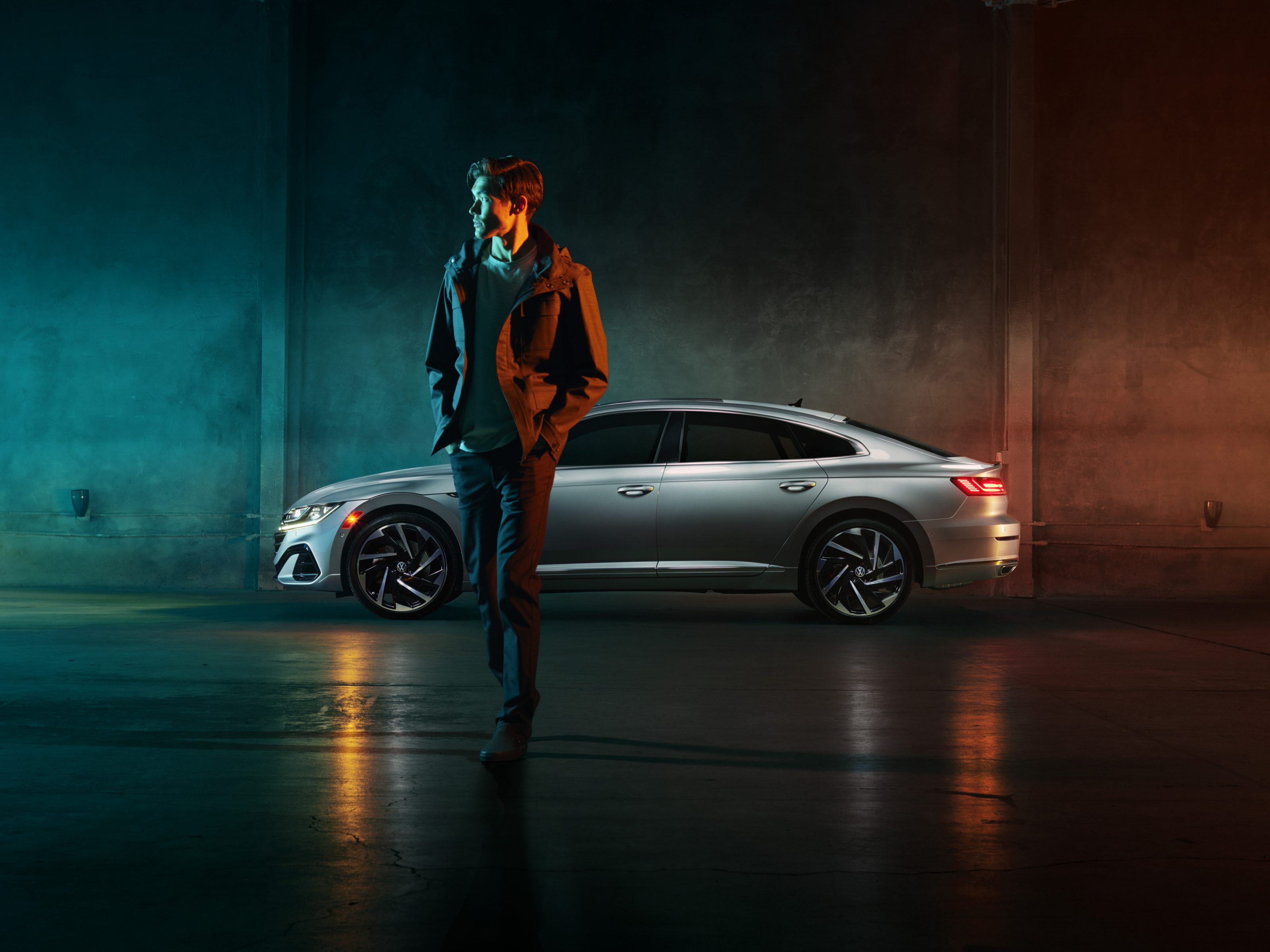 A man in a jacket stands in front of a silver car, surrounded by futuristic colors and lighting reminiscent of drone light painting, in a dimly lit indoor setting.