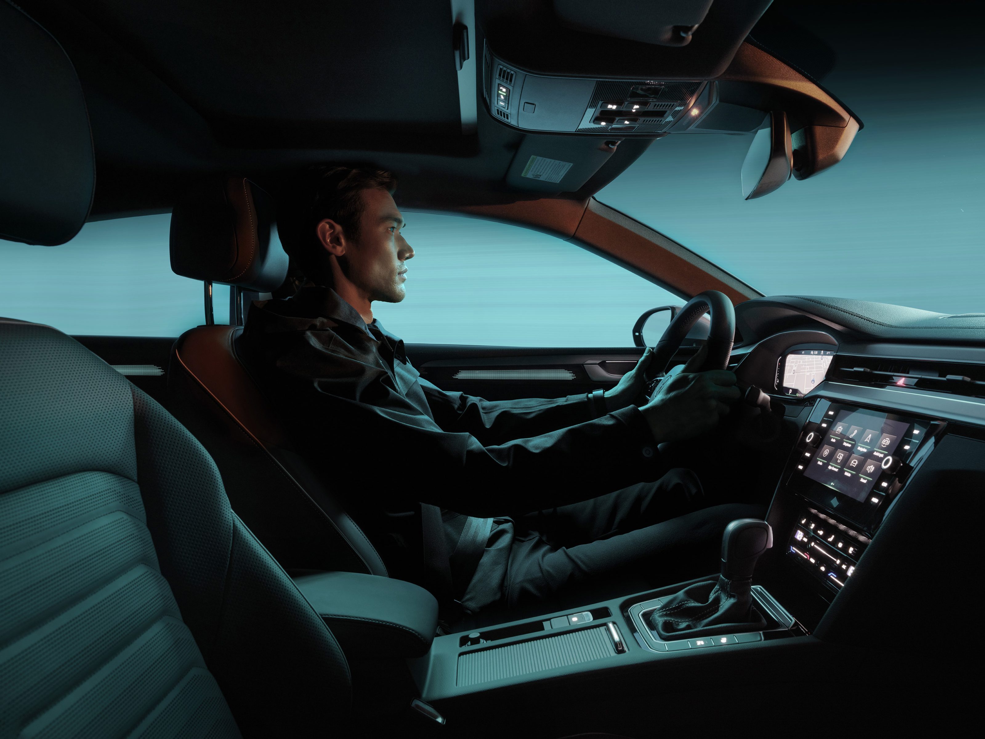 A man sits in the driver’s seat of a modern, futuristic car, hands on the wheel, with teal lighting casting an artistic glow inside.