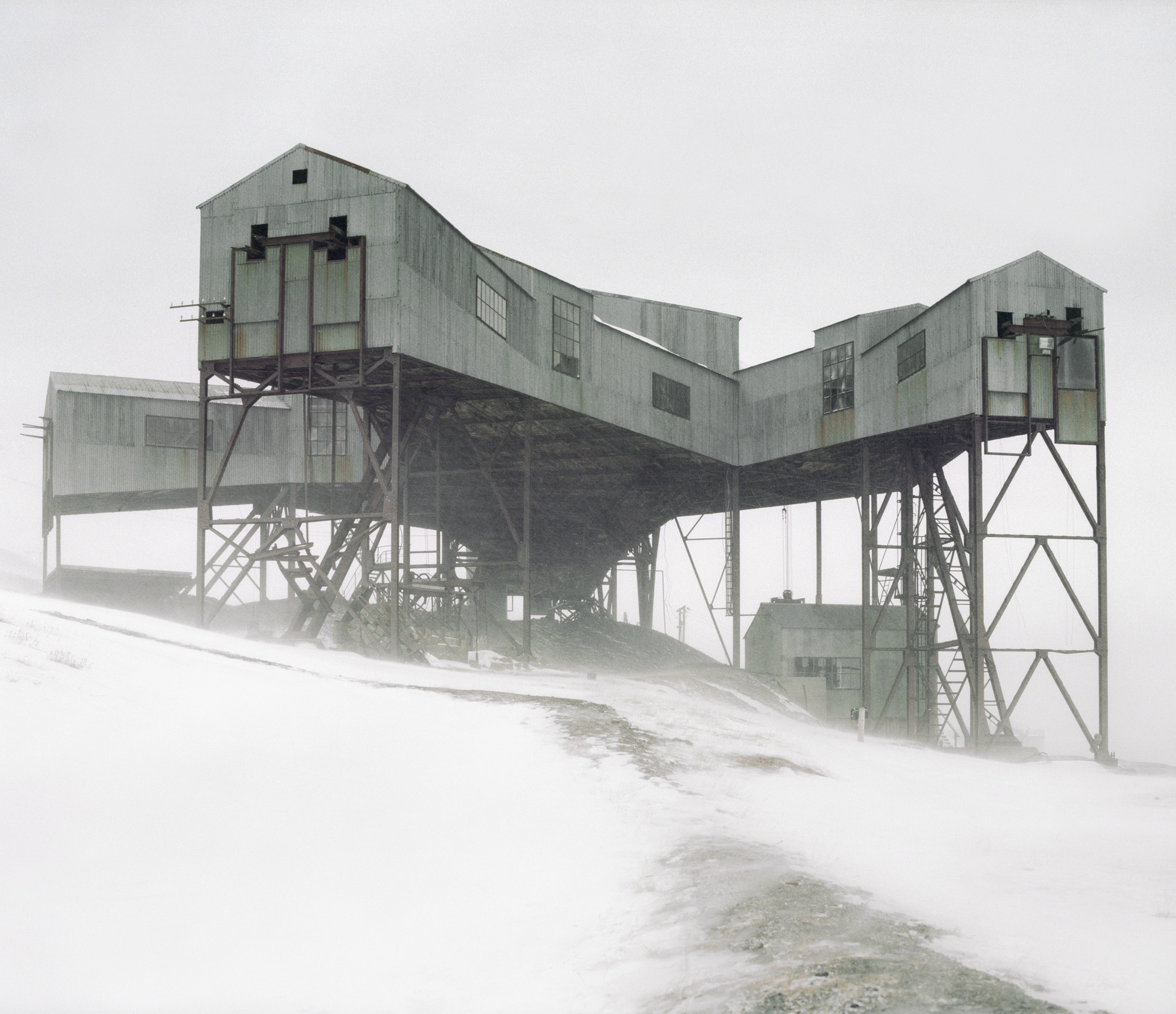 Large, weathered industrial structure on tall stilts stands over a snowy, misty landscape, reminiscent of a fine artist’s nighttime landscape photography.