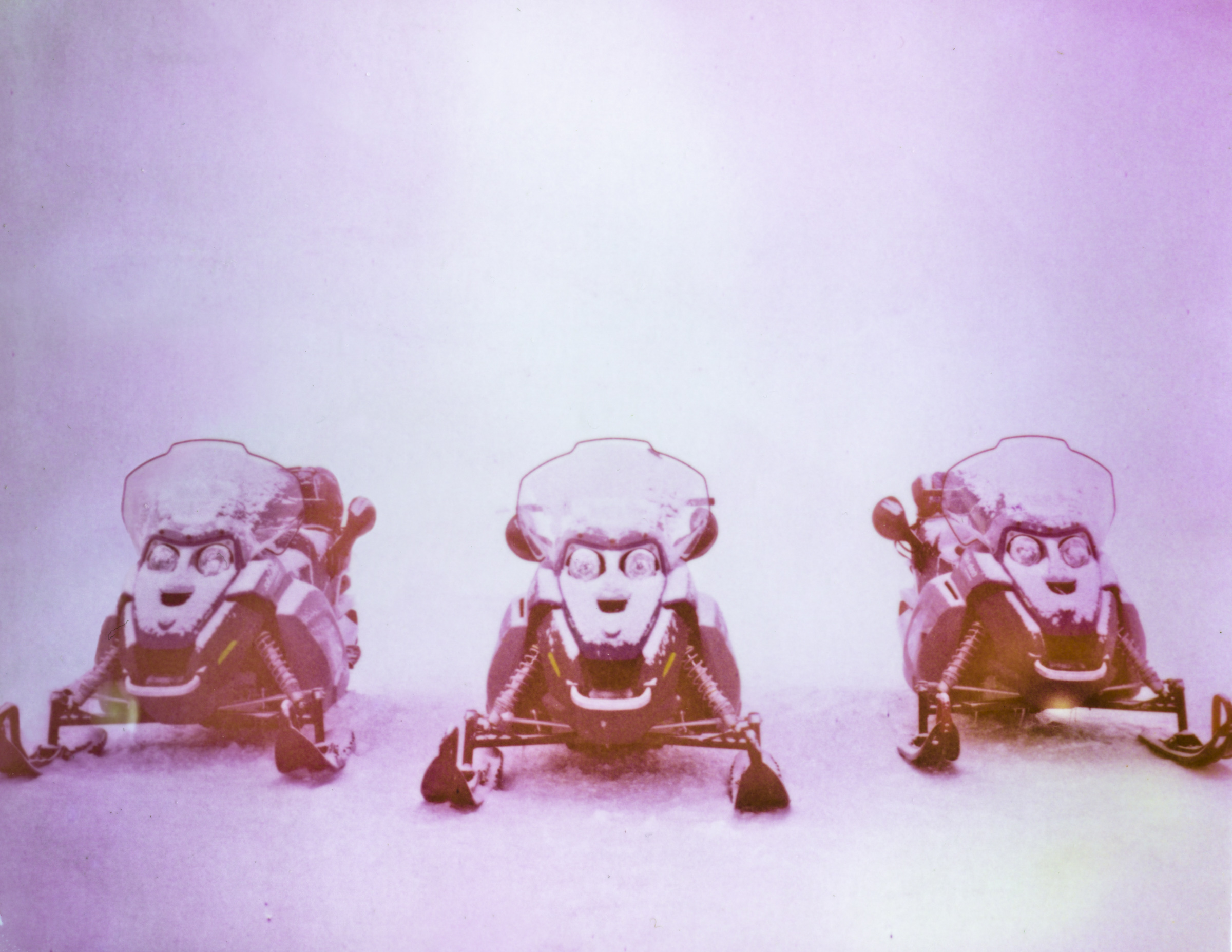 Three snowmobiles covered in snow are parked side by side on a snowy surface with a pink tint, capturing a futuristic vibe perfect for nighttime landscape photography.