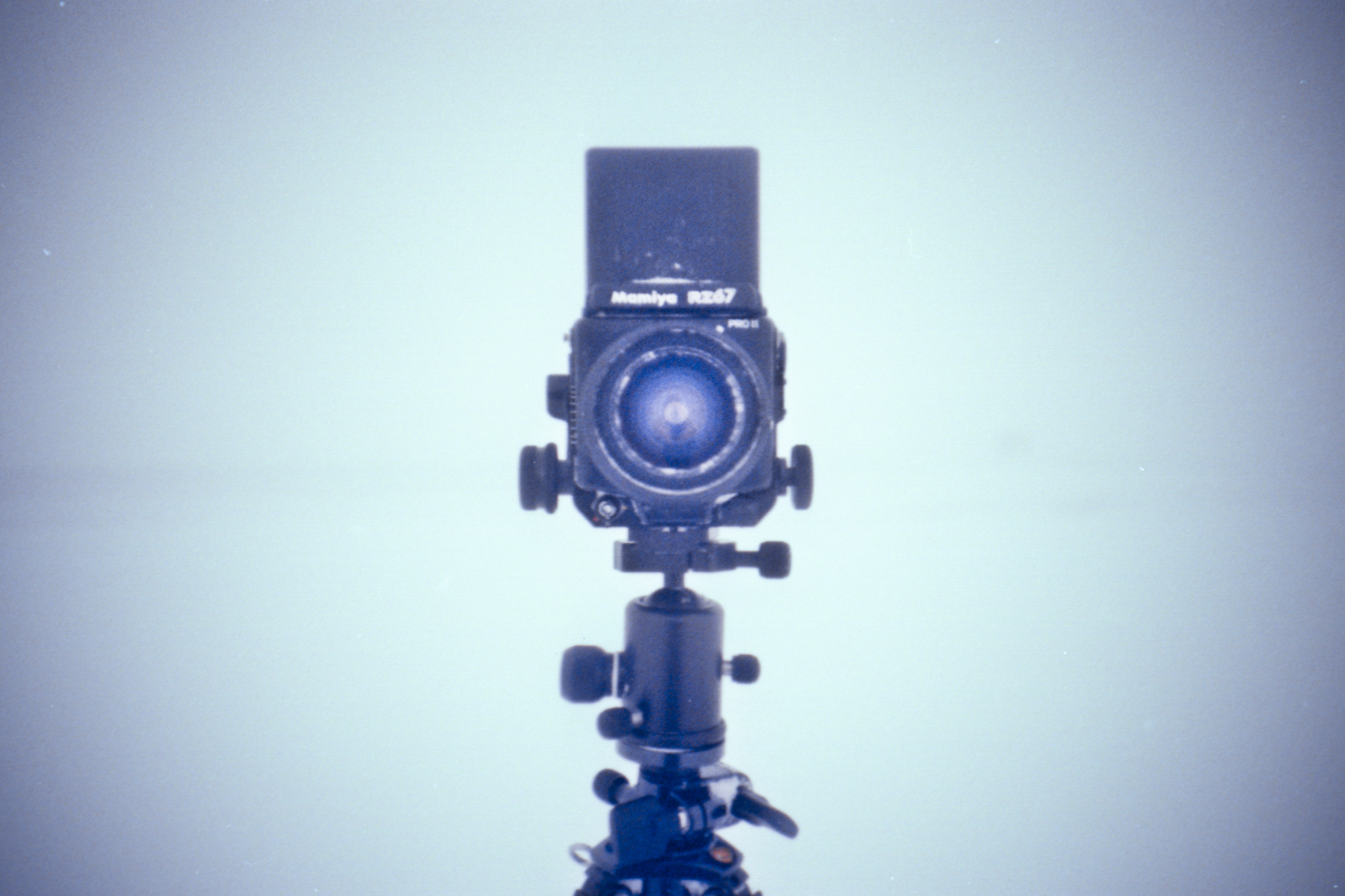 A vintage Mamiya RB67 camera on a tripod stands ready for a photographer against a plain, light blue background.