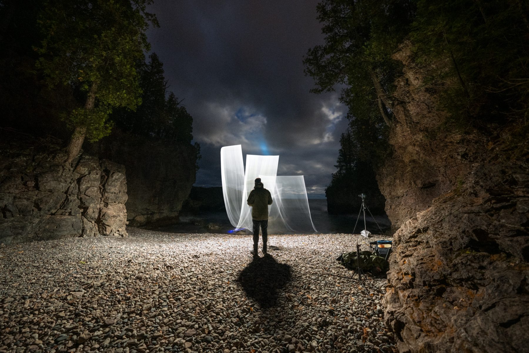 A photographer stands on rocky ground at night, capturing mesmerizing drone light art as trails of futuristic light weave between tall cliffs and trees.